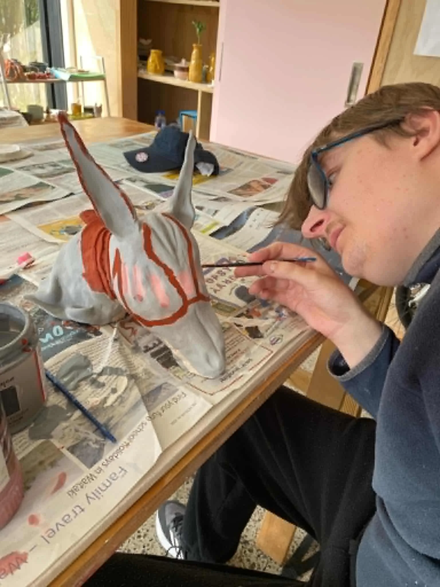 Louis painting a unicorn sculpture at a table covered with newspapers. Louis is wearing glasses and using a brush to paint details on the sculpture, which is still unfired clay white with orange markings.