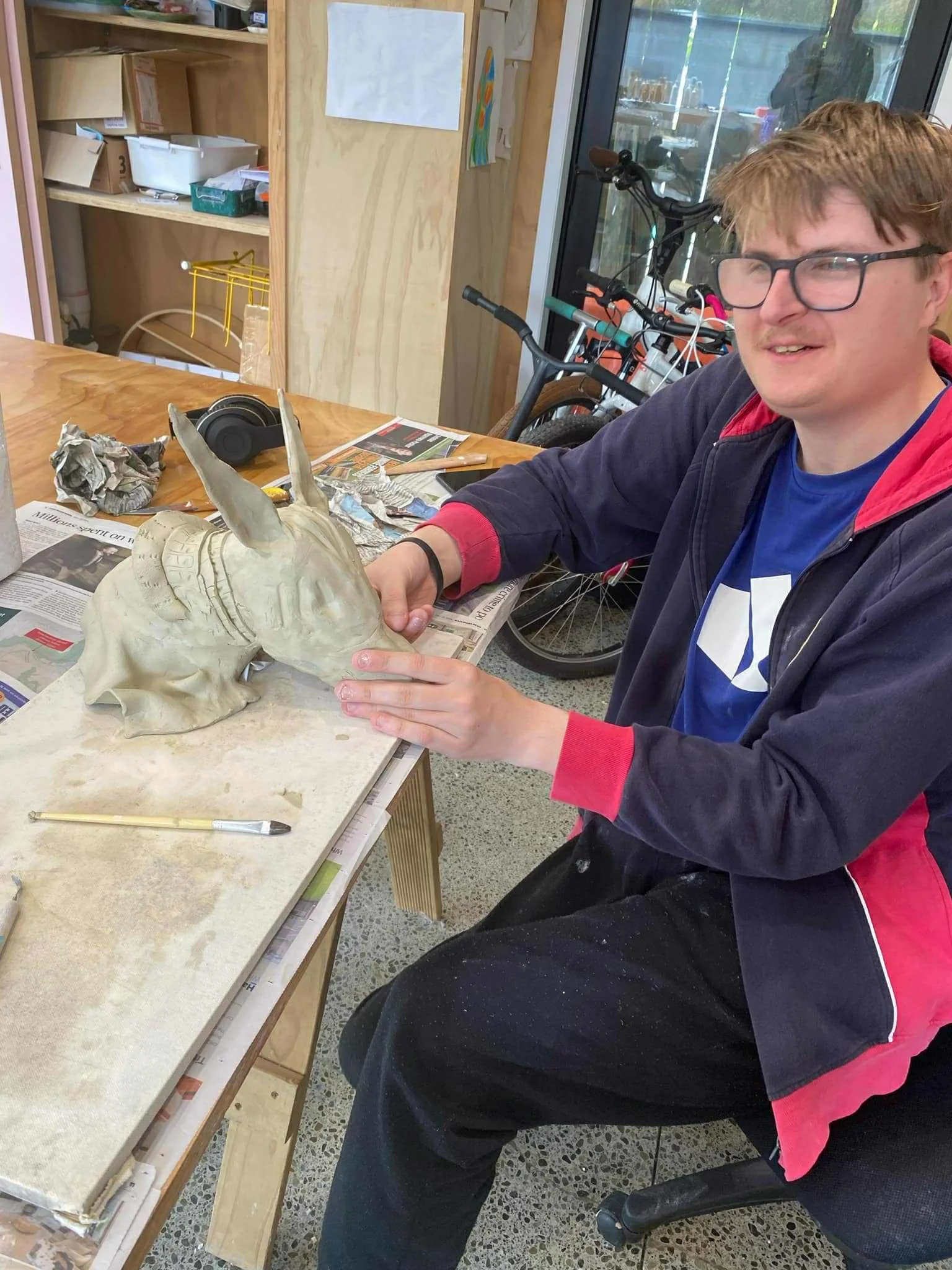 Louis with glasses is sitting at a work table, holding a ceramic sculpture with upright ears. The table has tools and newspapers on it, and there are bicycles and shelves in the background.