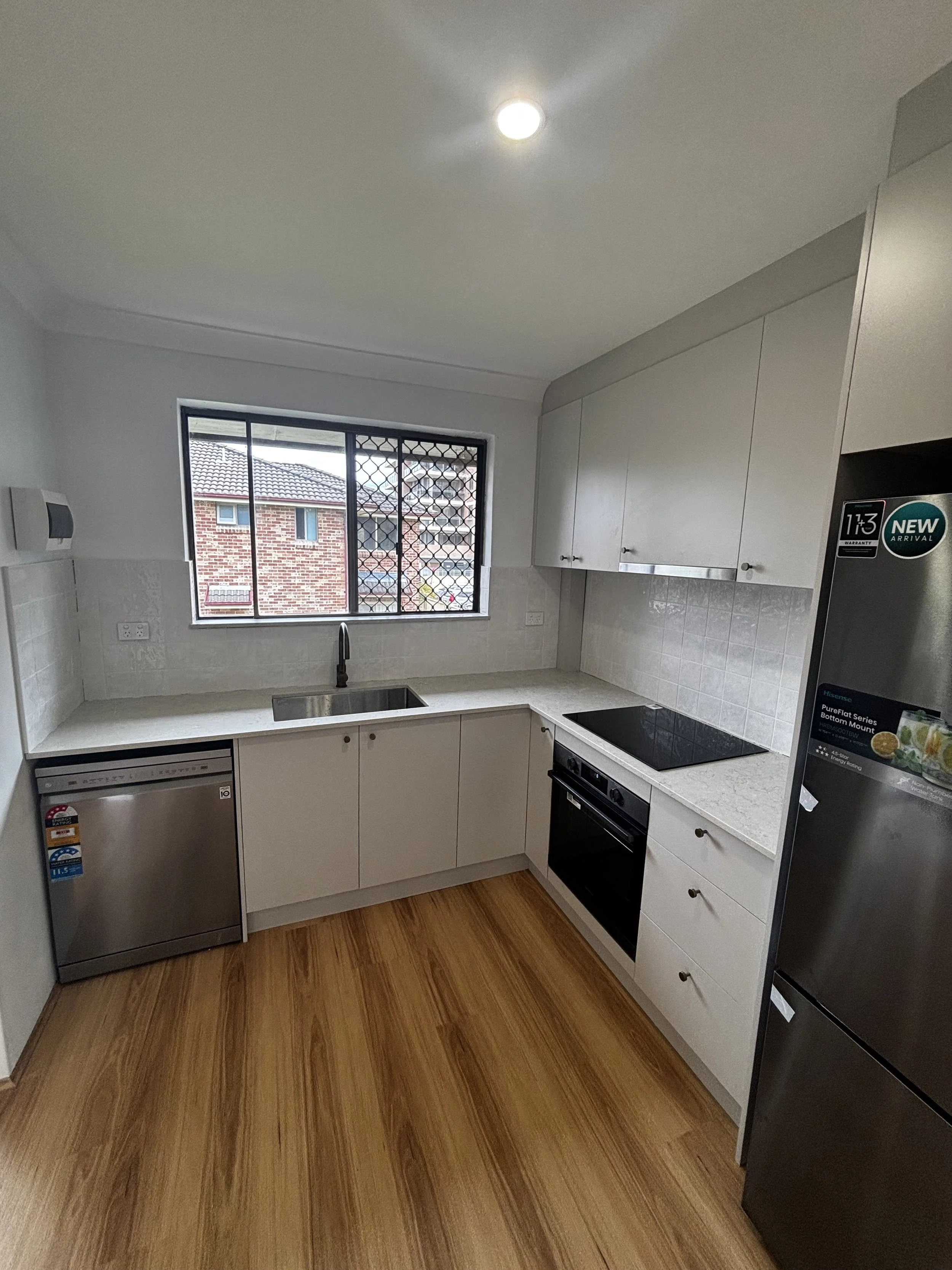 Modern kitchen with white cabinets, a window with security bars, a stainless steel dishwasher, a black electric stove, and a refrigerator with stickers, all on a wooden floor.