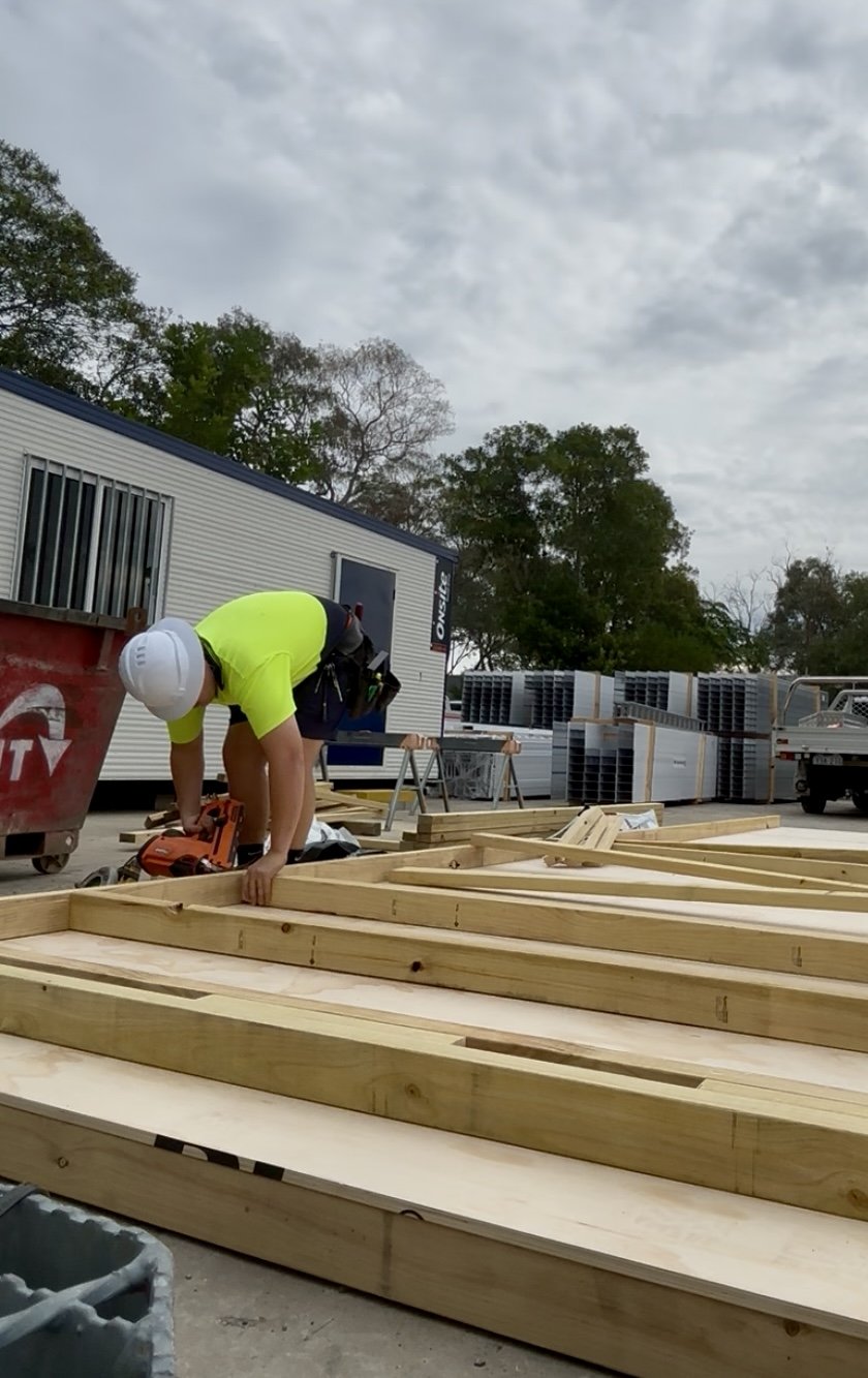 A construction worker wearing a white helmet and a bright yellow shirt is working with wooden beams on a building site. The worker is kneeling and using a power tool amid construction materials and equipment, with a trailer, trees, and an overcast sky in the background.