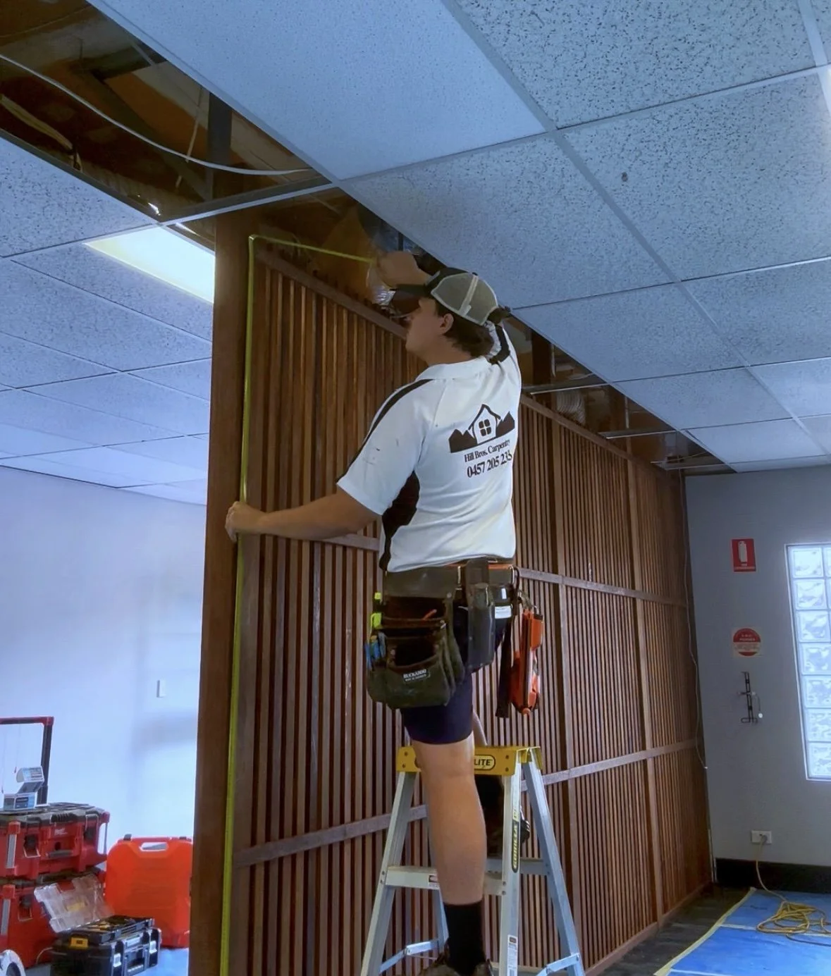 A worker on a step ladder measuring a wooden wall installation in a room with a dropped ceiling and tools on the floor.
