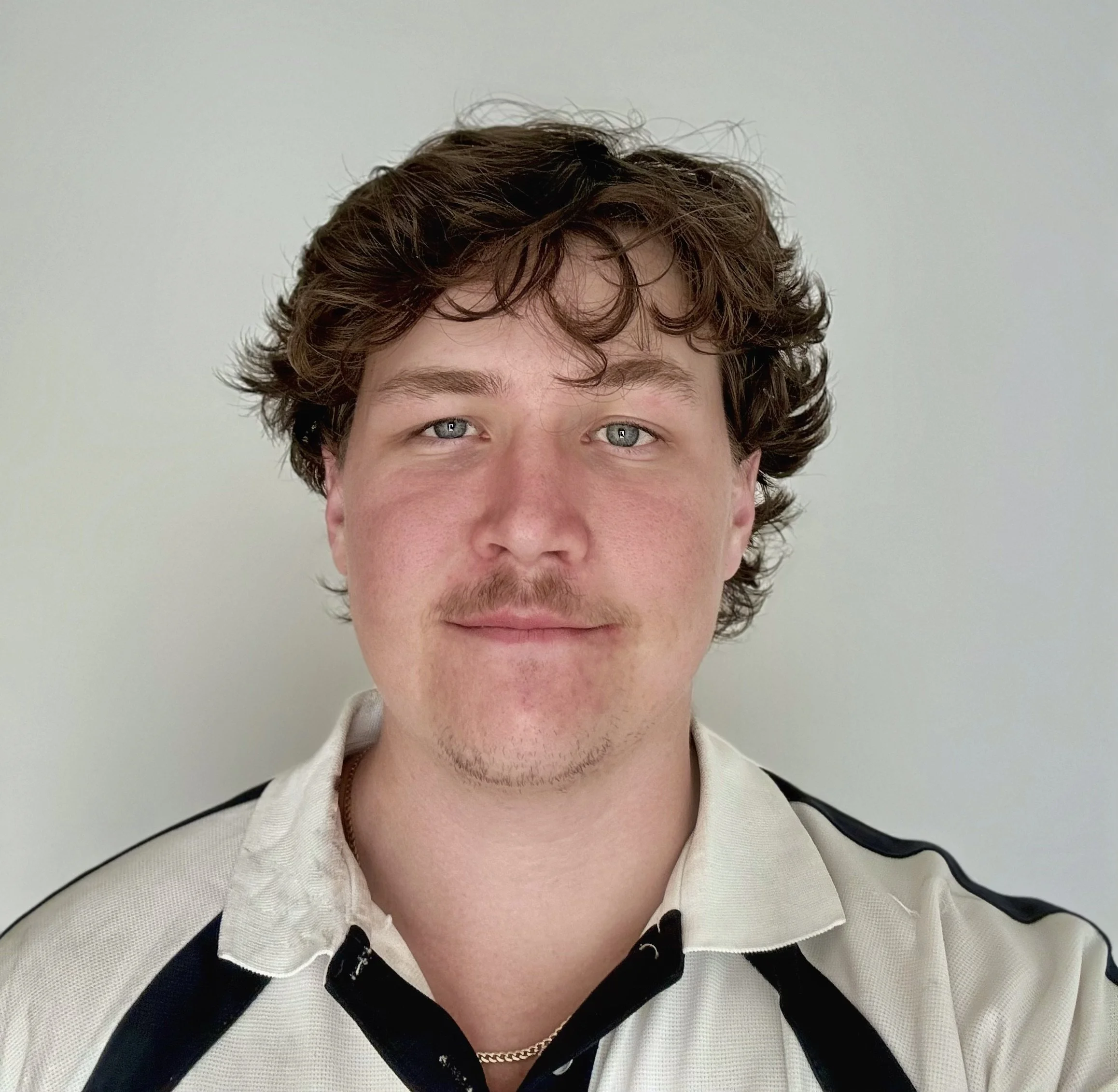Close-up of a young man with curly brown hair, blue eyes, and light skin, wearing a beige and black striped shirt and a gold chain, against a plain light background.