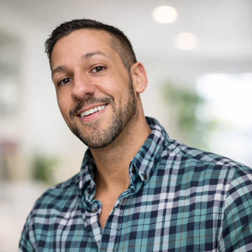 Close-up of a smiling man with short brown hair and a beard, wearing a blue plaid shirt, indoors with a blurred background.