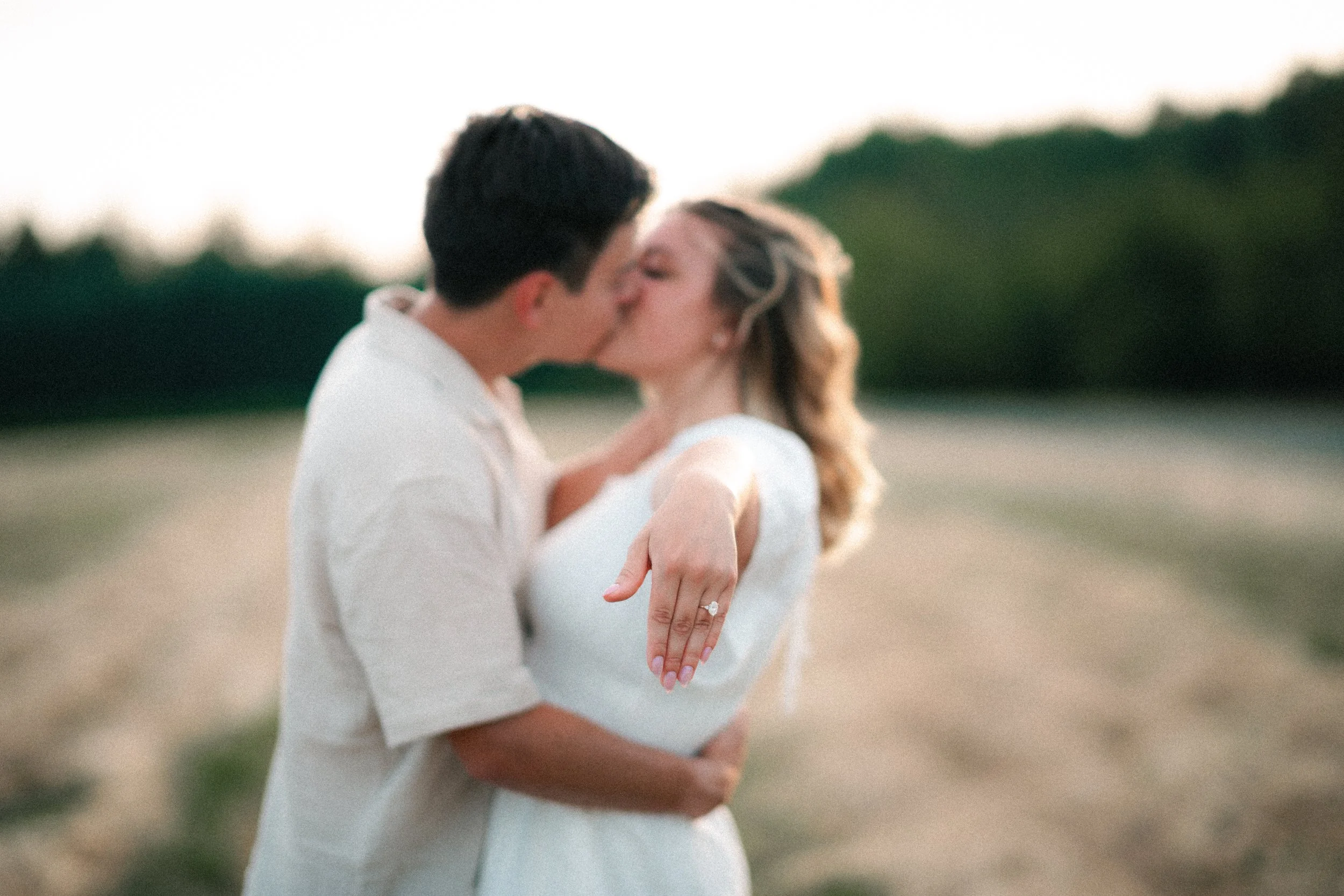 A couple sharing a kiss outdoors, with the woman pointing at her wedding ring, on a path with trees in the background.