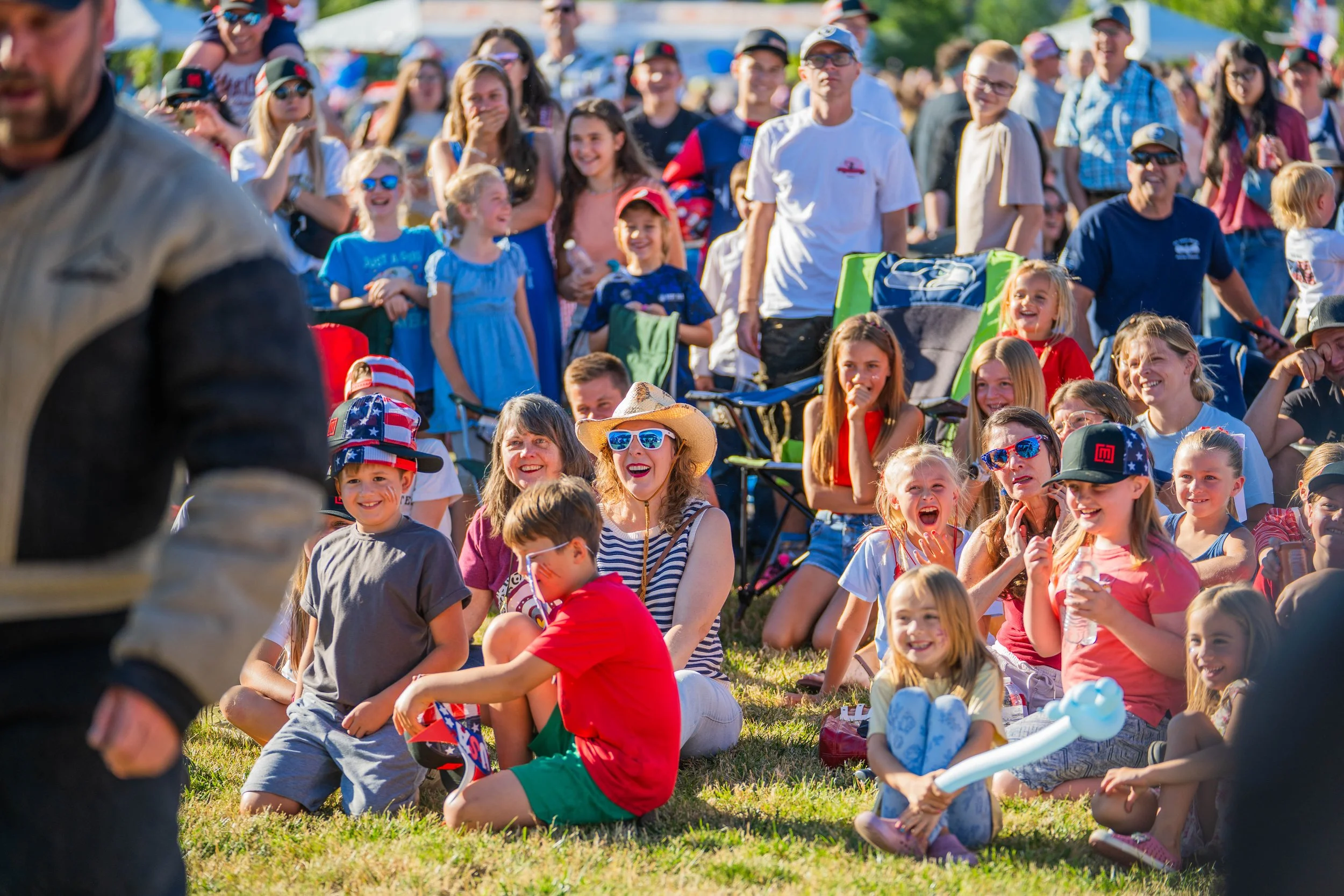 A large crowd of children and adults gathered outside on a sunny day, sitting on the grass, attending an event. The children are dressed in red, white, and blue clothing, some wearing patriotic hats. The crowd appears cheerful and engaged, with some 