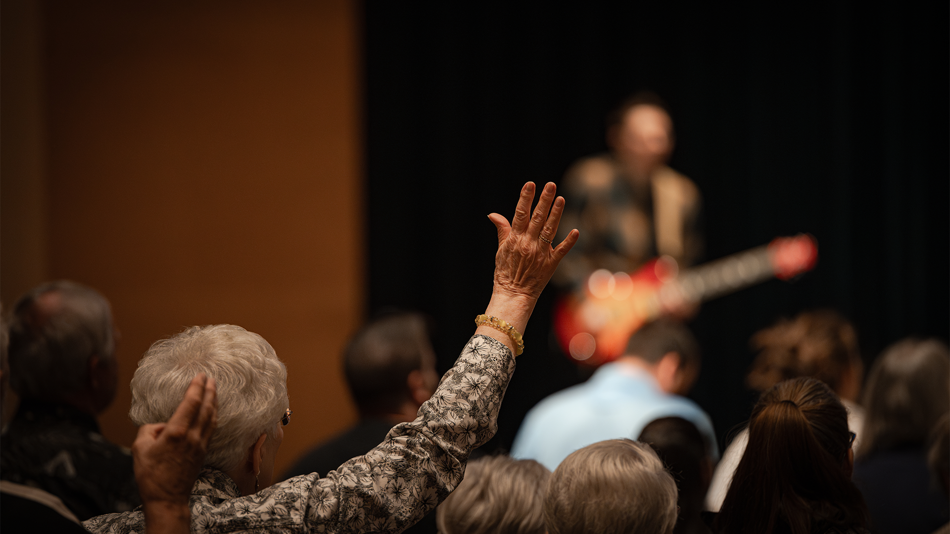 Audience members raise their hands during a music or speech event, with a performing musician visible in the background on stage.