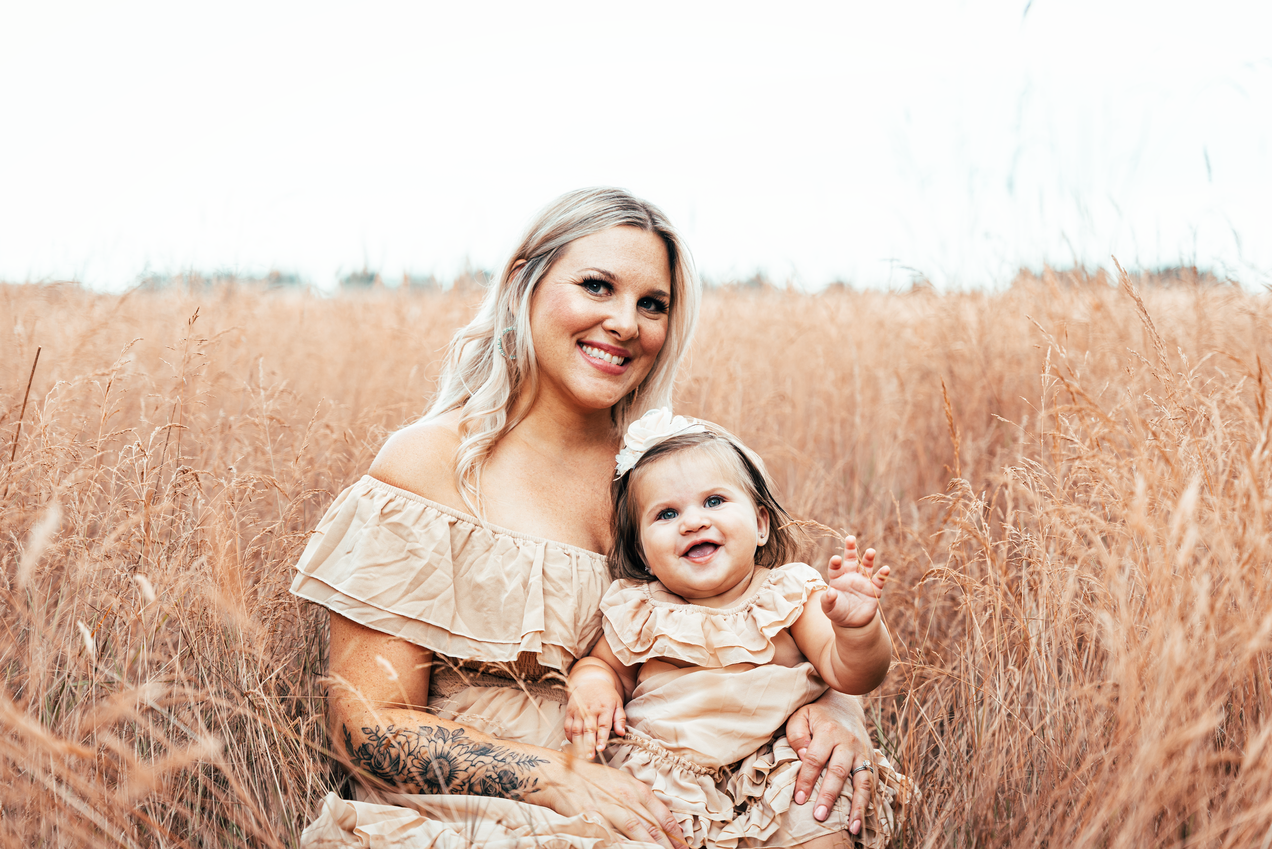 A woman with blonde hair and tattoo sleeve holding a smiling toddler with gray hair in a beige dress in a field of tall, golden grass under a bright, lightly clouded sky.