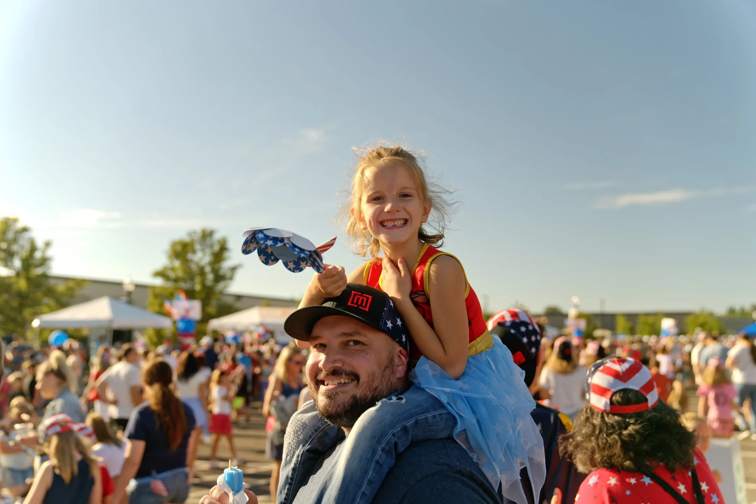 A girl smiling and holding a pinwheel sitting on a man's shoulders at an outdoor event with a large crowd in the background.