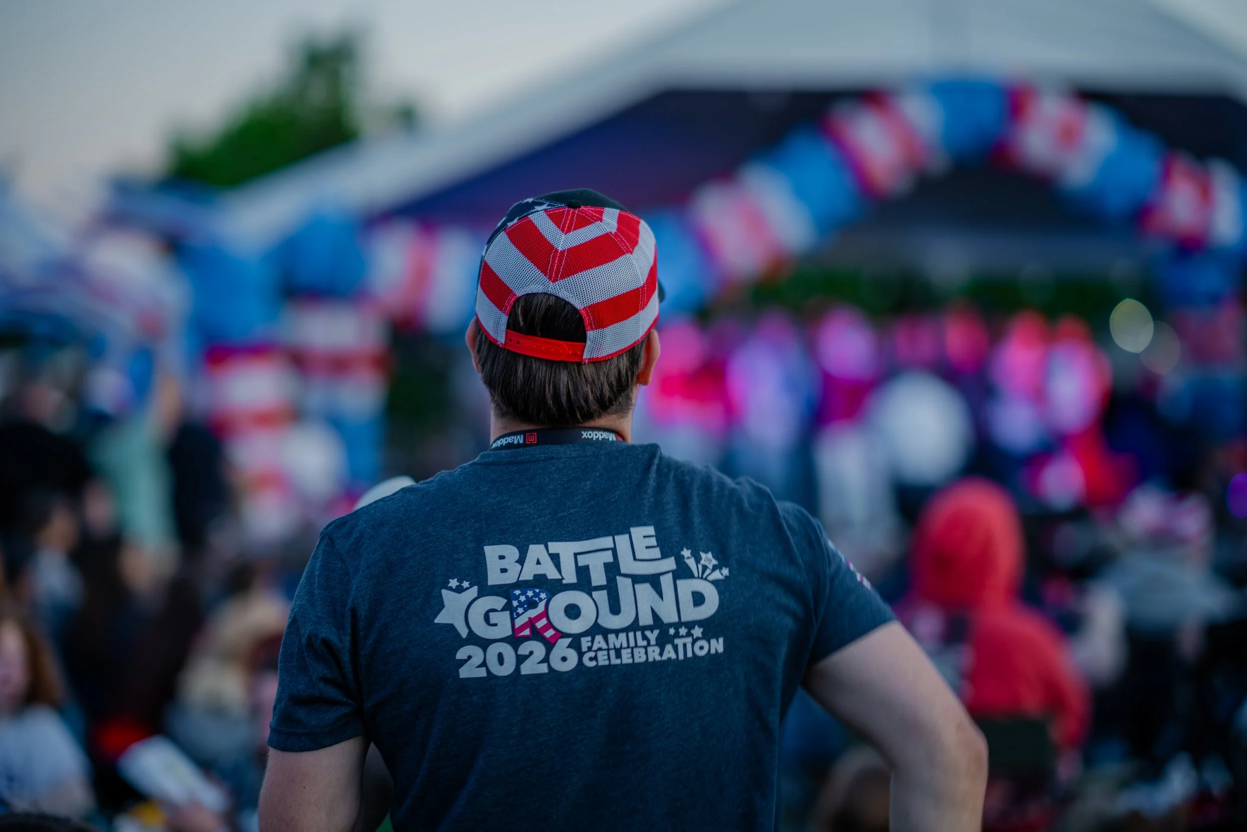 Young man wearing a patriotic red, white, and blue baseball cap and a dark blue t-shirt with the words 'Battle Ground' and '2026' on the back, attending an outdoor event with colorful flags and banners in the background.