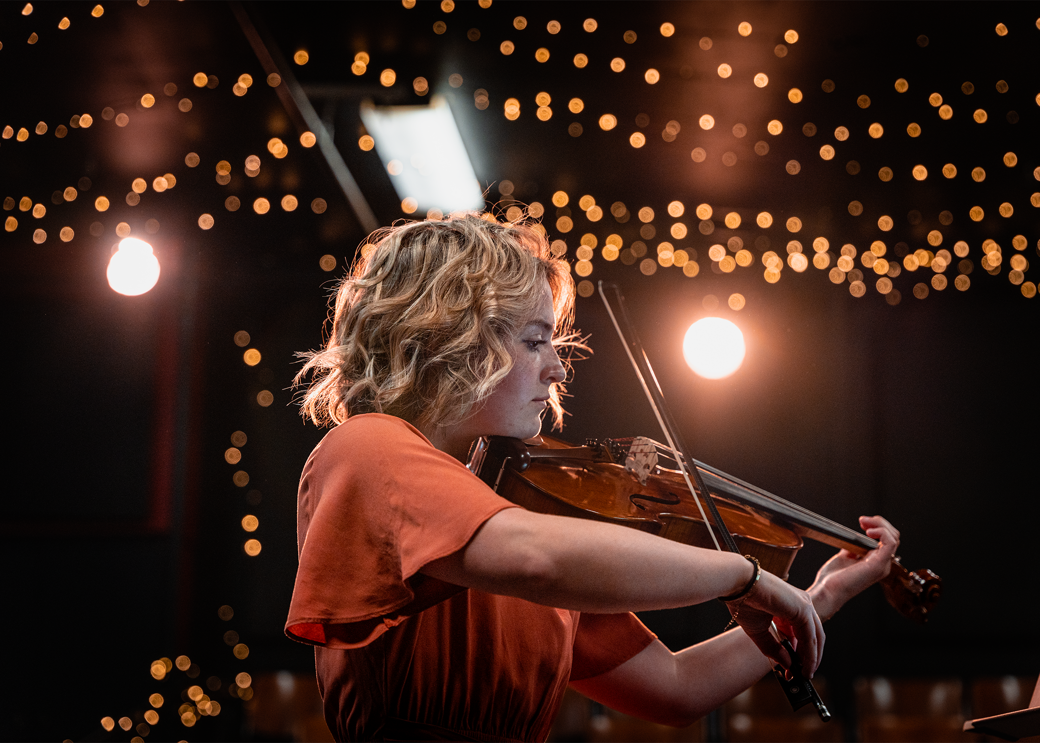 A woman with curly blonde hair playing the violin on a stage or indoor setting with a background of warm string lights.