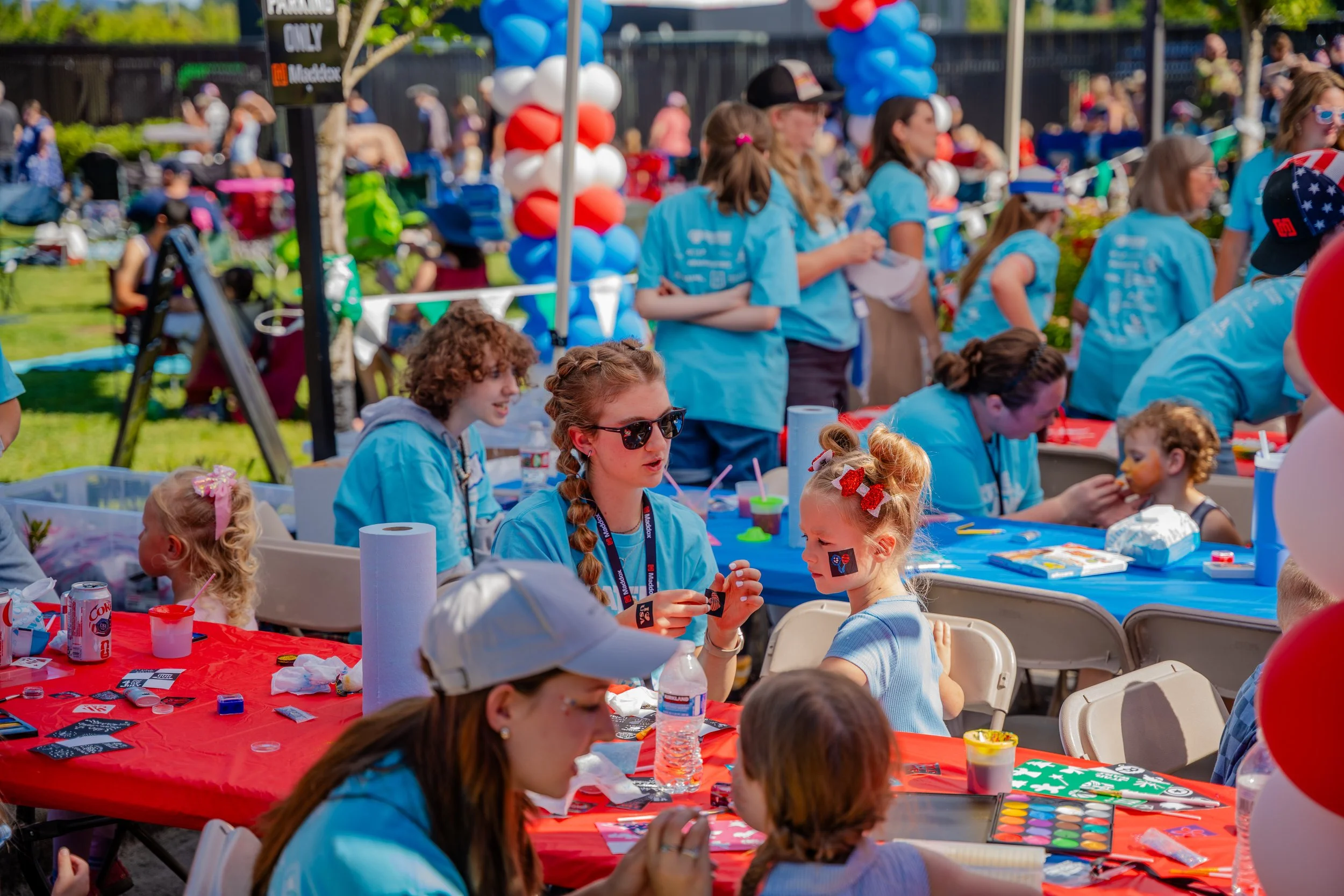 Children and adults participating in an outdoor festive event with tables decorated in red, white, and blue. Some children have face paint and are engaging in arts and crafts activities.