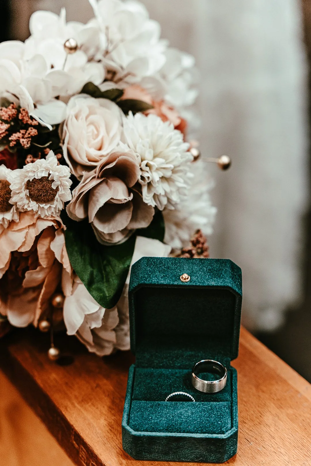 A bouquet of light-colored artificial flowers, including roses and hydrangeas, next to an open green velvet ring box containing a silver wedding band, placed on a wooden surface.