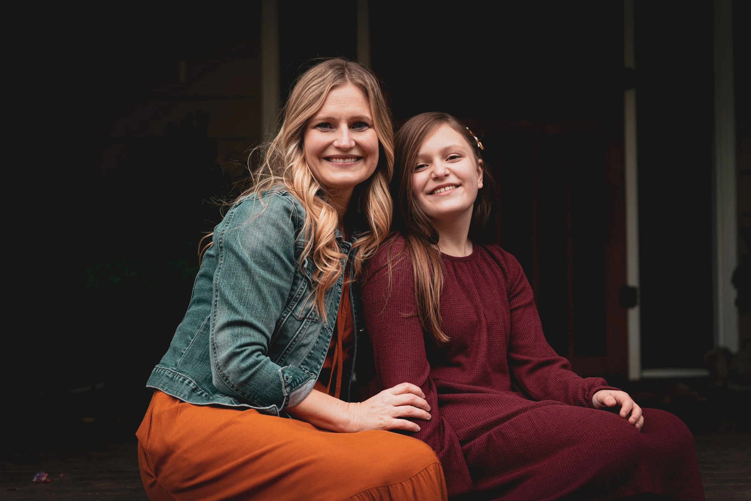 A woman with long blonde hair and a girl with long brown hair sitting close together and smiling, outdoors at night.
