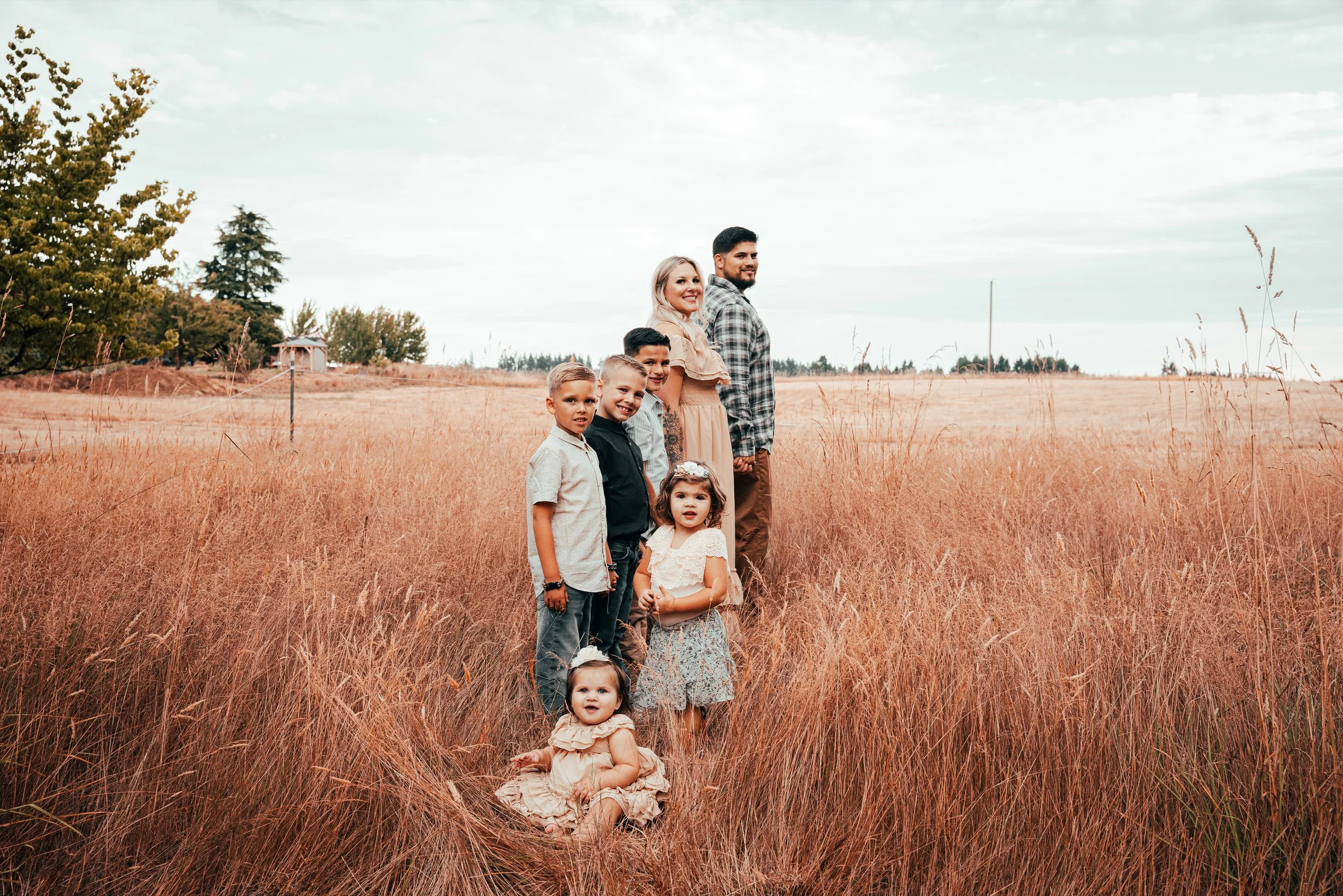 A family of seven stands in a field of tall, orange-brown grass with trees and a cloudy sky in the background. The family includes three children, two women, and two men, all smiling and holding hands.