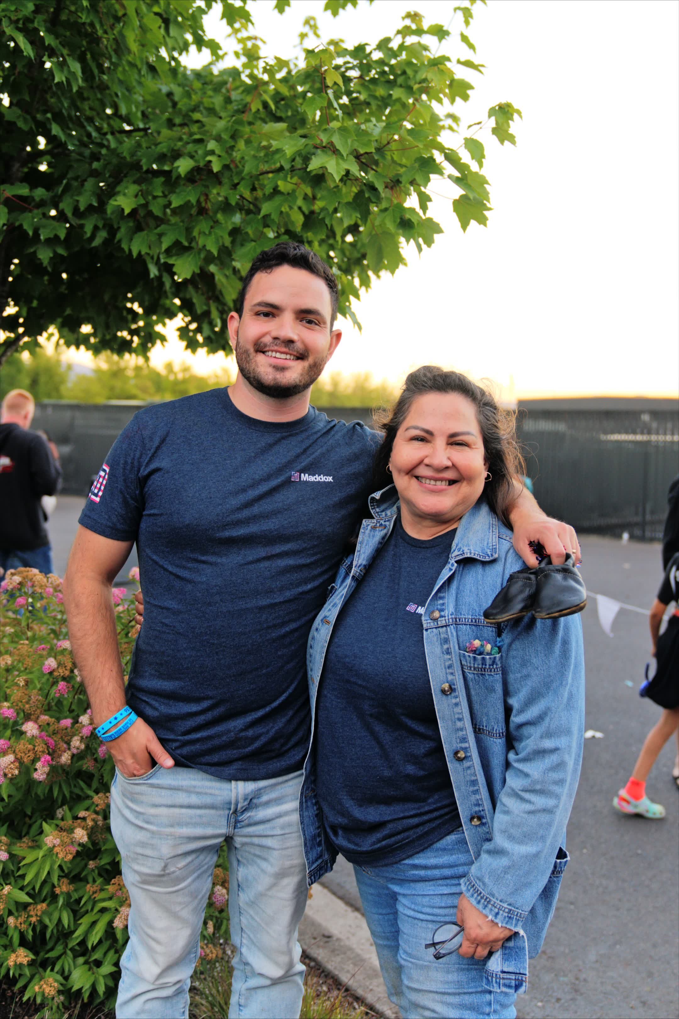 A man and woman smiling and standing close together outdoors, with a green tree and other people in the background during sunset.