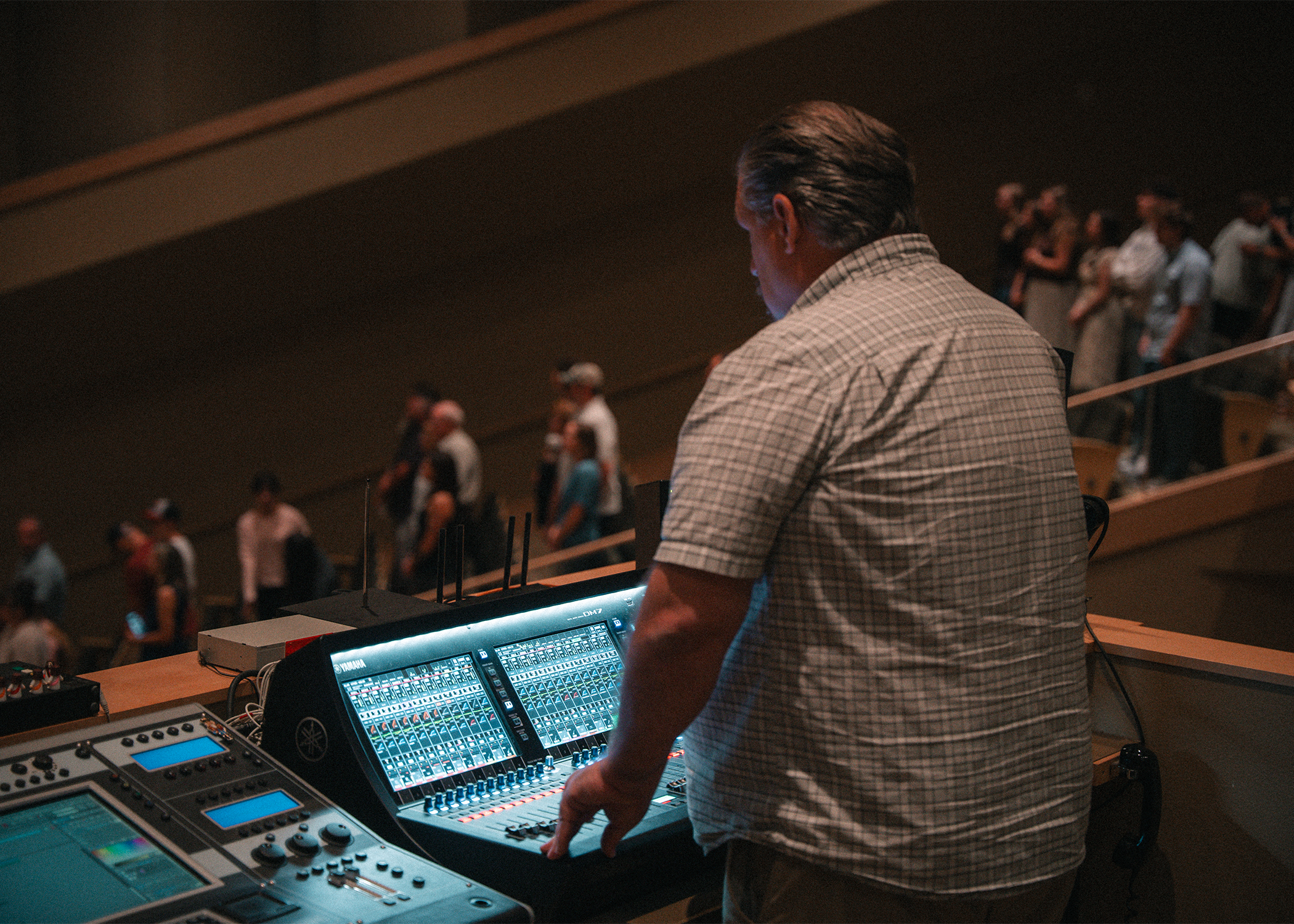 A person with gray hair wearing a beige checkered shirt operating a digital music mixer with multiple screen controls in a dark auditorium.