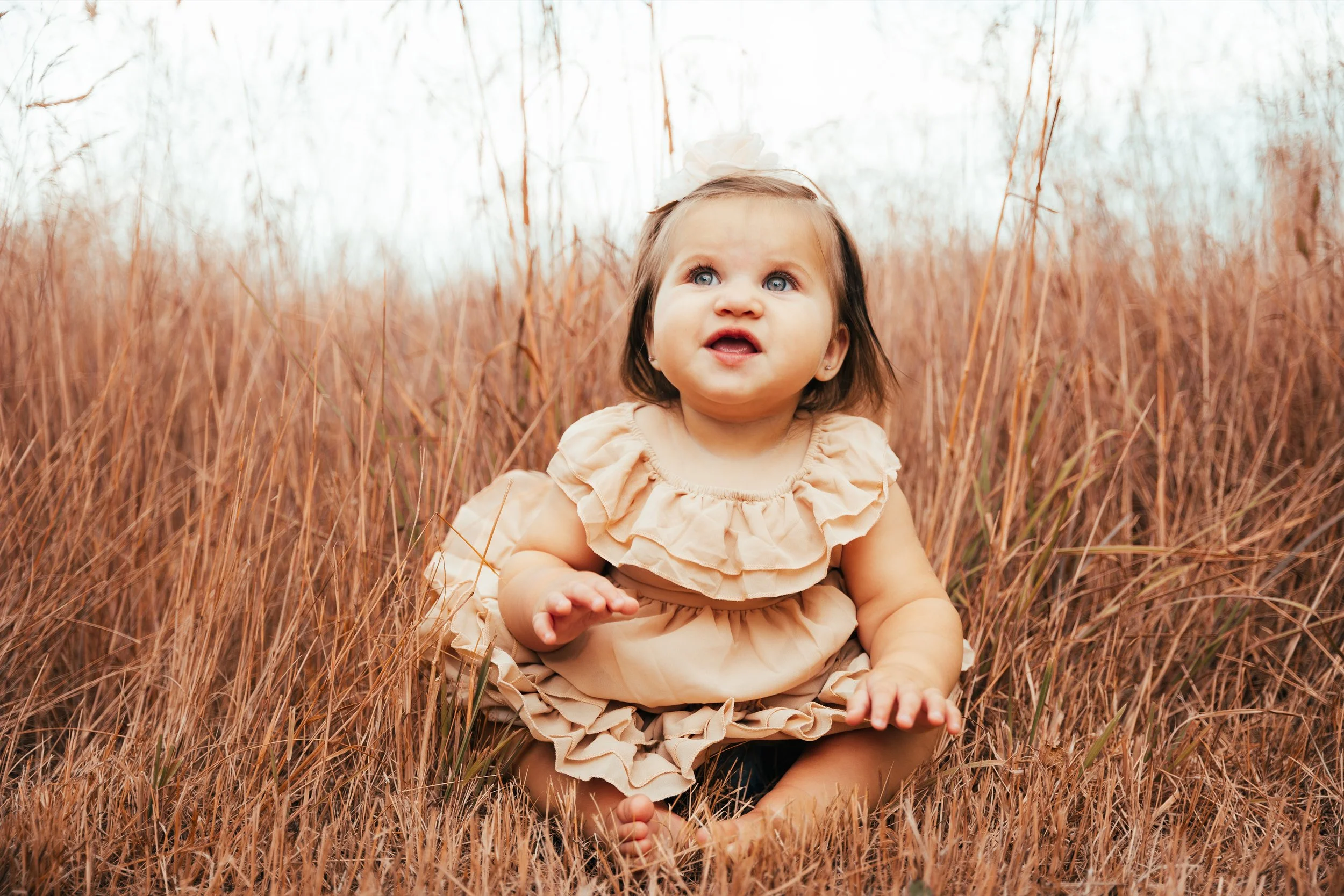 A young girl with blue eyes and brown hair, wearing a beige dress with ruffles, sitting in tall, golden-brown grass outdoors on a slightly overcast day.