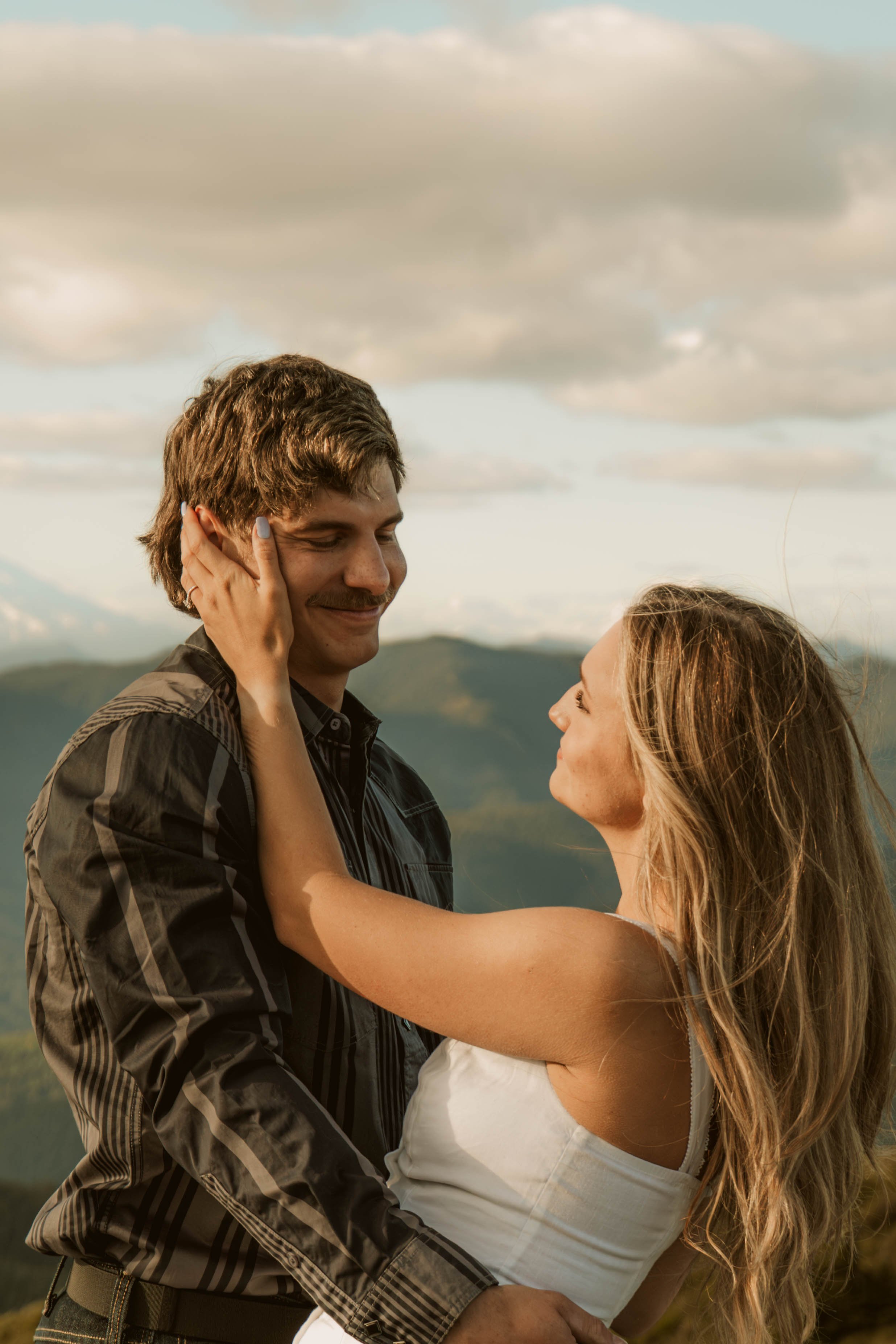 A man and woman embrace outdoors, looking at each other with a scenic landscape and sky in the background.