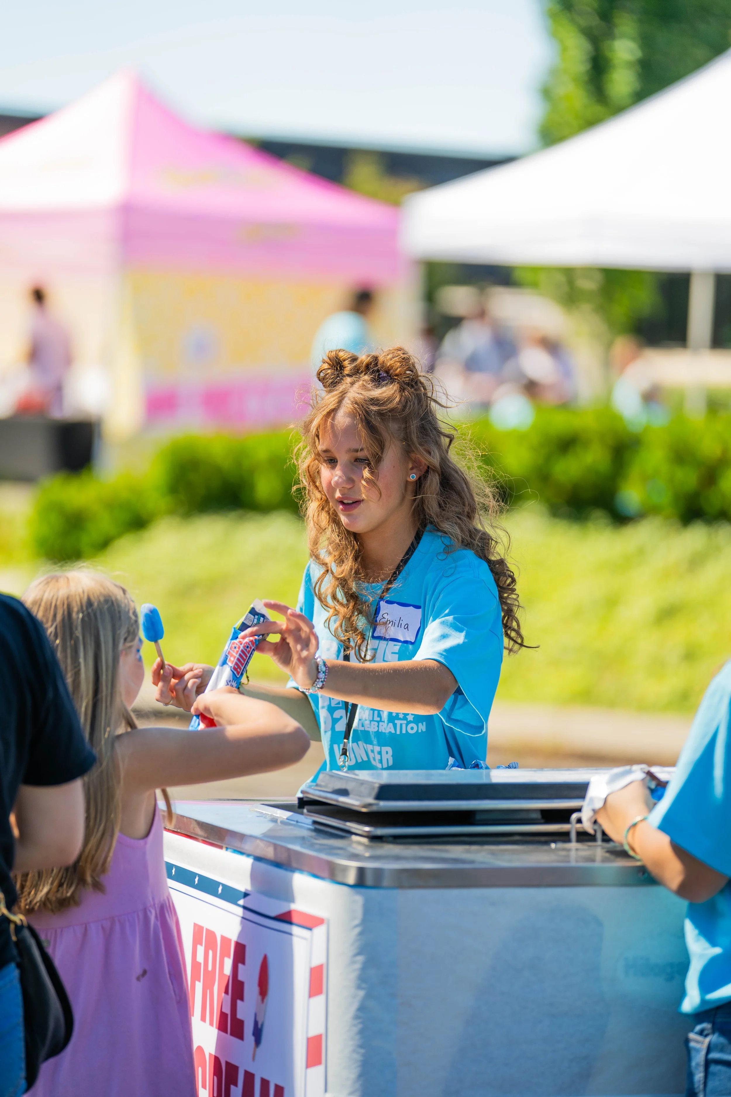 A young girl with curly hair, wearing a blue shirt, is handing a popsicle to a girl in a pink dress at a community event with tents and people in the background.