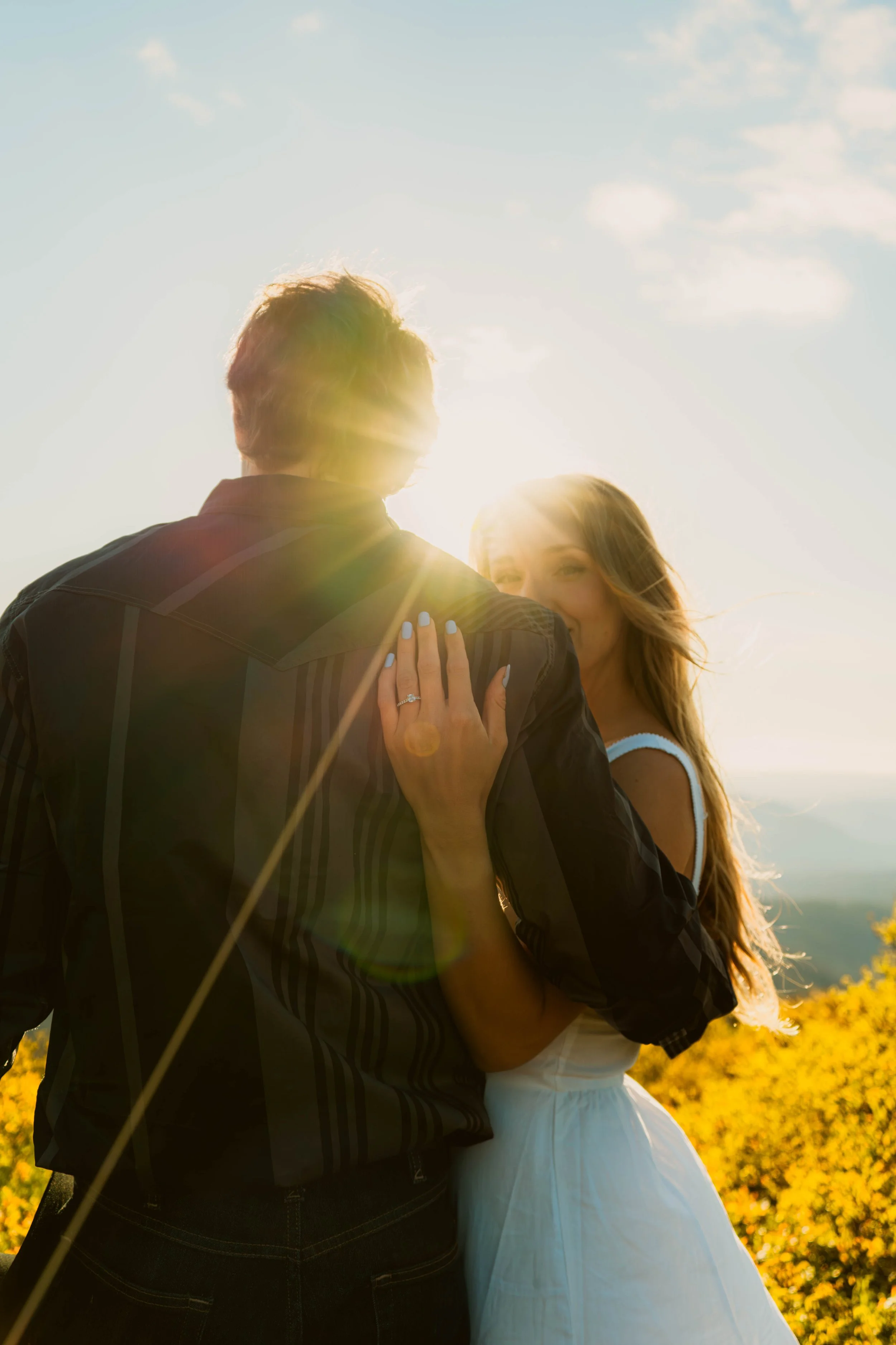 A couple standing outdoors, with the man facing away and the woman smiling, during sunset with sunlight creating a lens flare, in a natural setting with trees and a blue sky with a few clouds.
