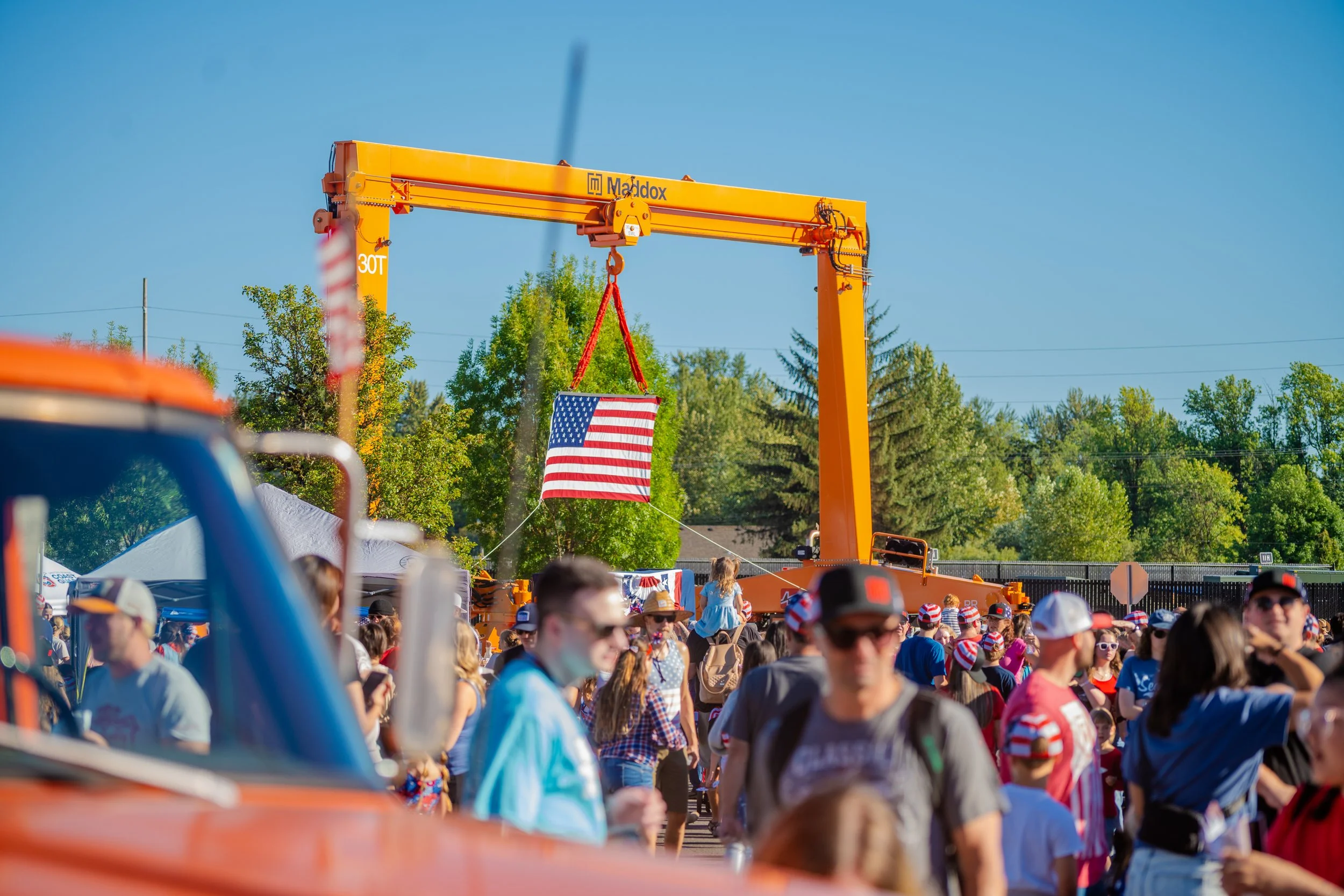 A crowd gathered outdoors during a sunny day, with a large orange construction crane displaying an American flag hanging from it in the background, and many people wearing patriotic red, white, and blue clothing and hats.