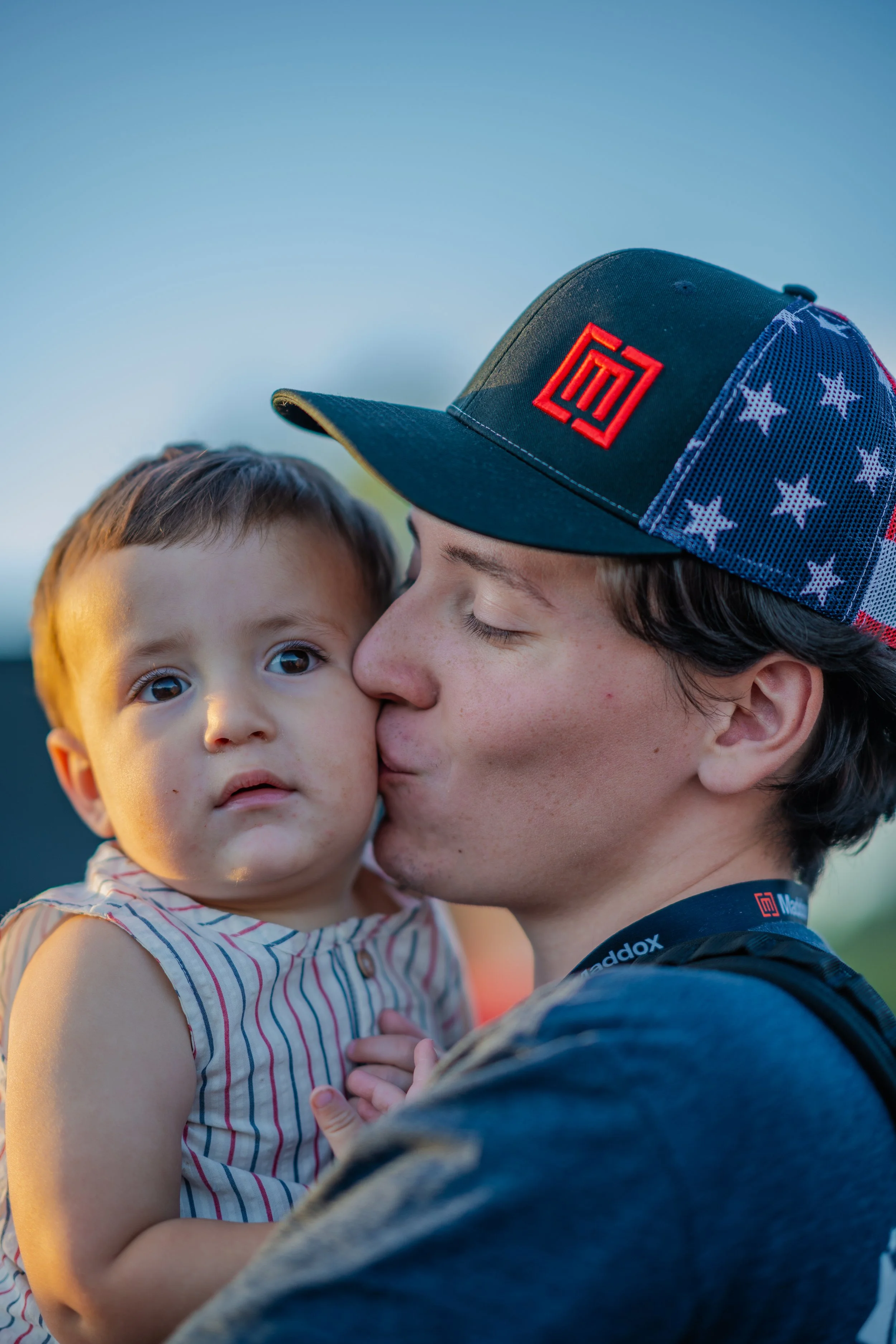 A young man wearing a cap with American flag design and red logo is holding and kissing a young girl with short hair and big eyes, wearing a striped sleeveless top, outdoors during sunset.