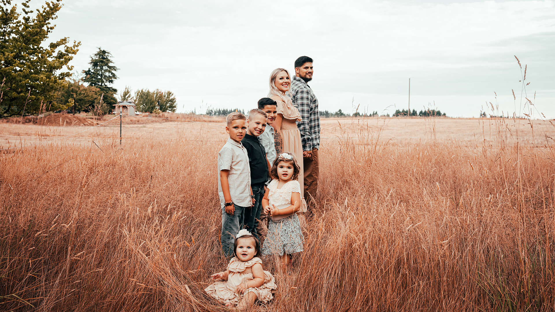 A family of nine standing in a field of tall, dry grass with a few trees and a gazebo in the background, under a cloudy sky.