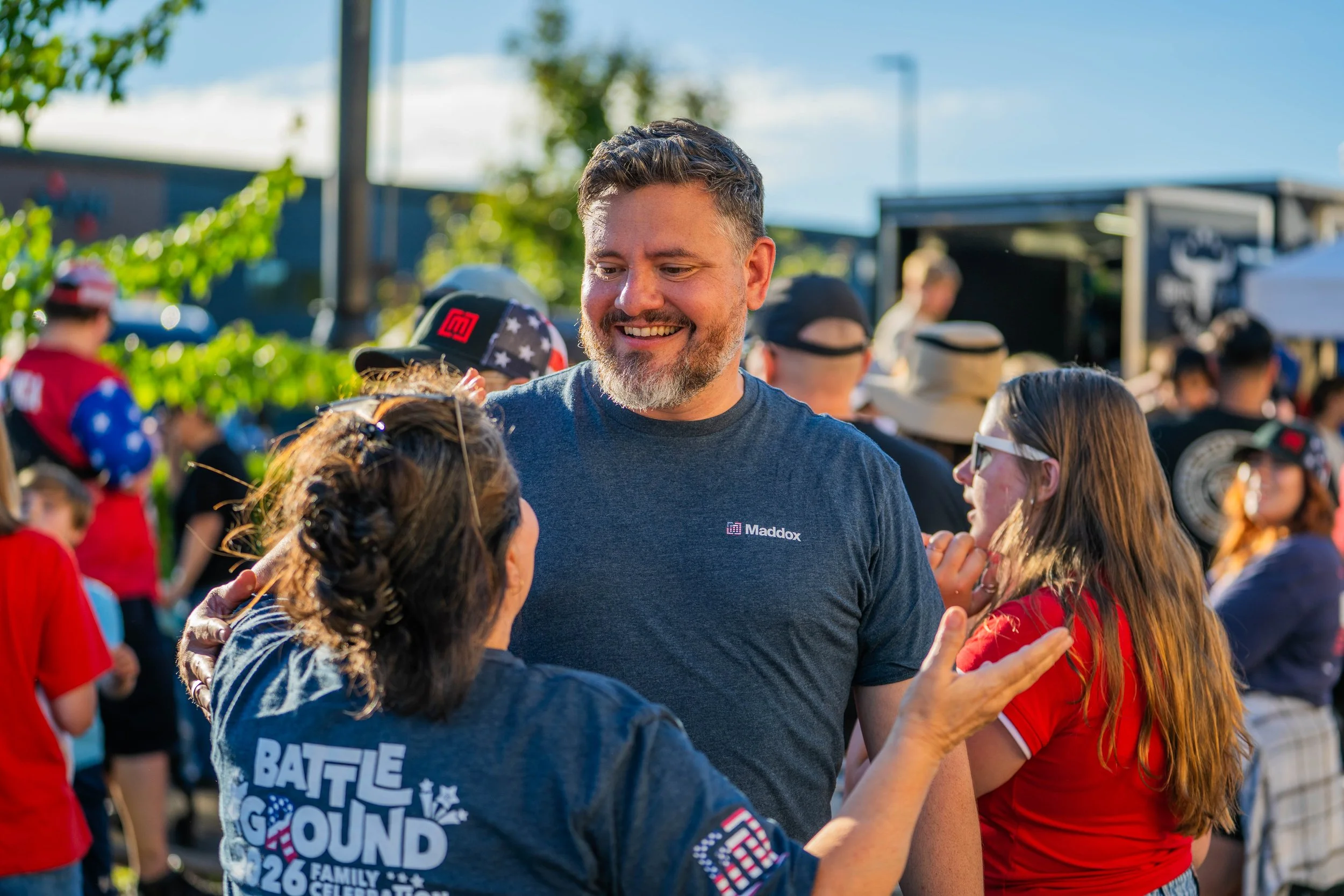 A man with short dark hair and a beard smiling and hugging a woman in a crowd outdoors during daytime. The man is wearing a blue T-shirt with white and red writing. The woman has glasses and braided hair. The background has many people, some with hat