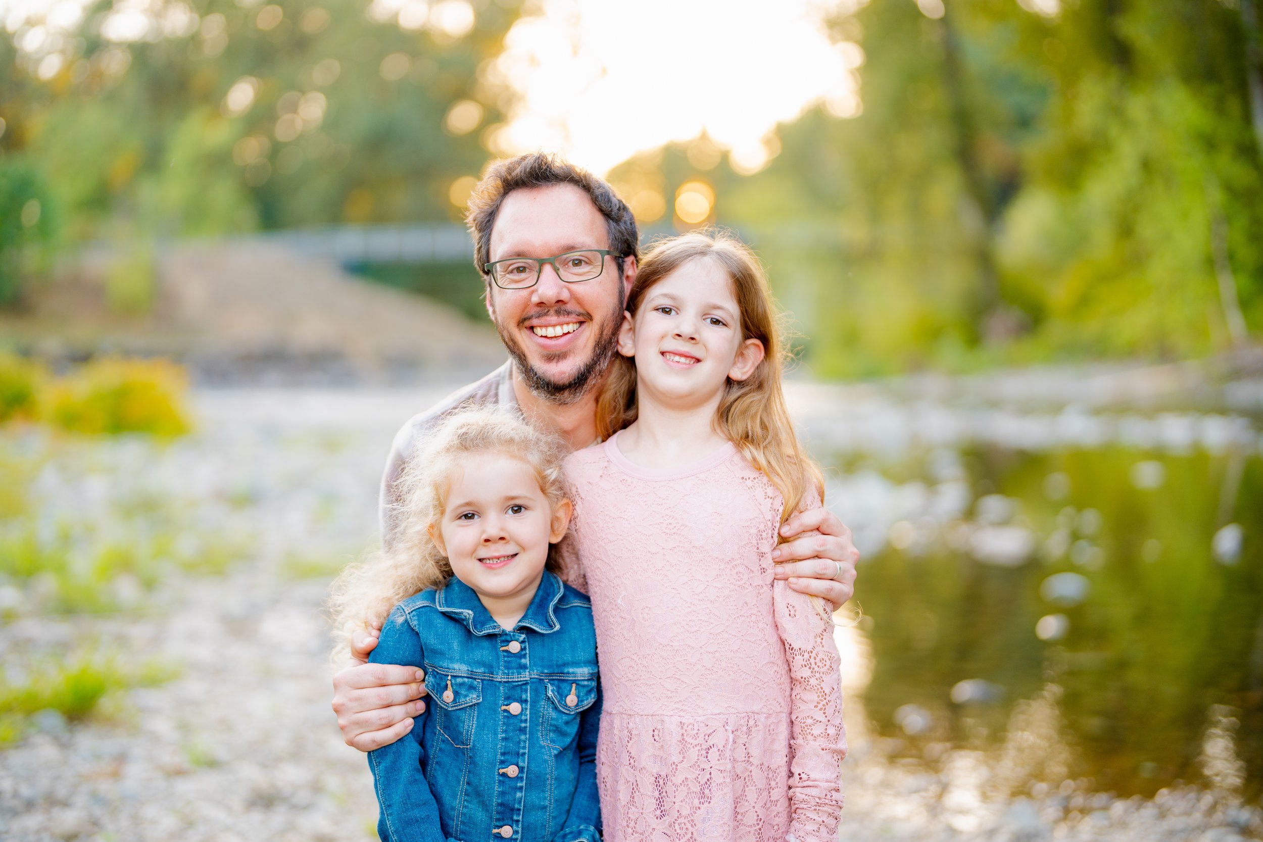 A man and three young girls are smiling outdoors near a river with green trees and sunlight in the background.