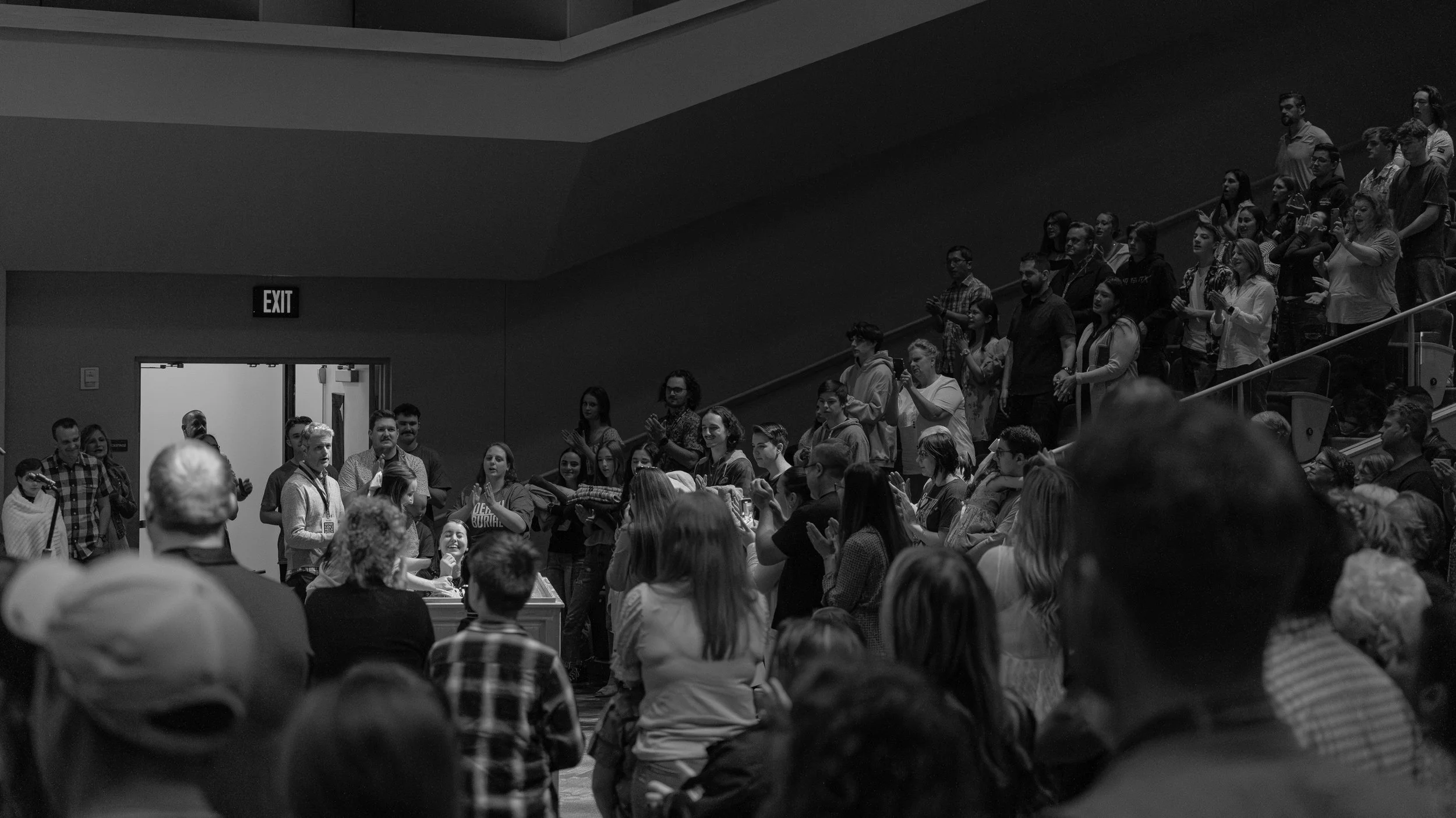 Crowd of people gathered in an auditorium, with some standing and clapping, and others sitting and watching. The setting appears to be a public event or gathering, with a diverse group of attendees.
