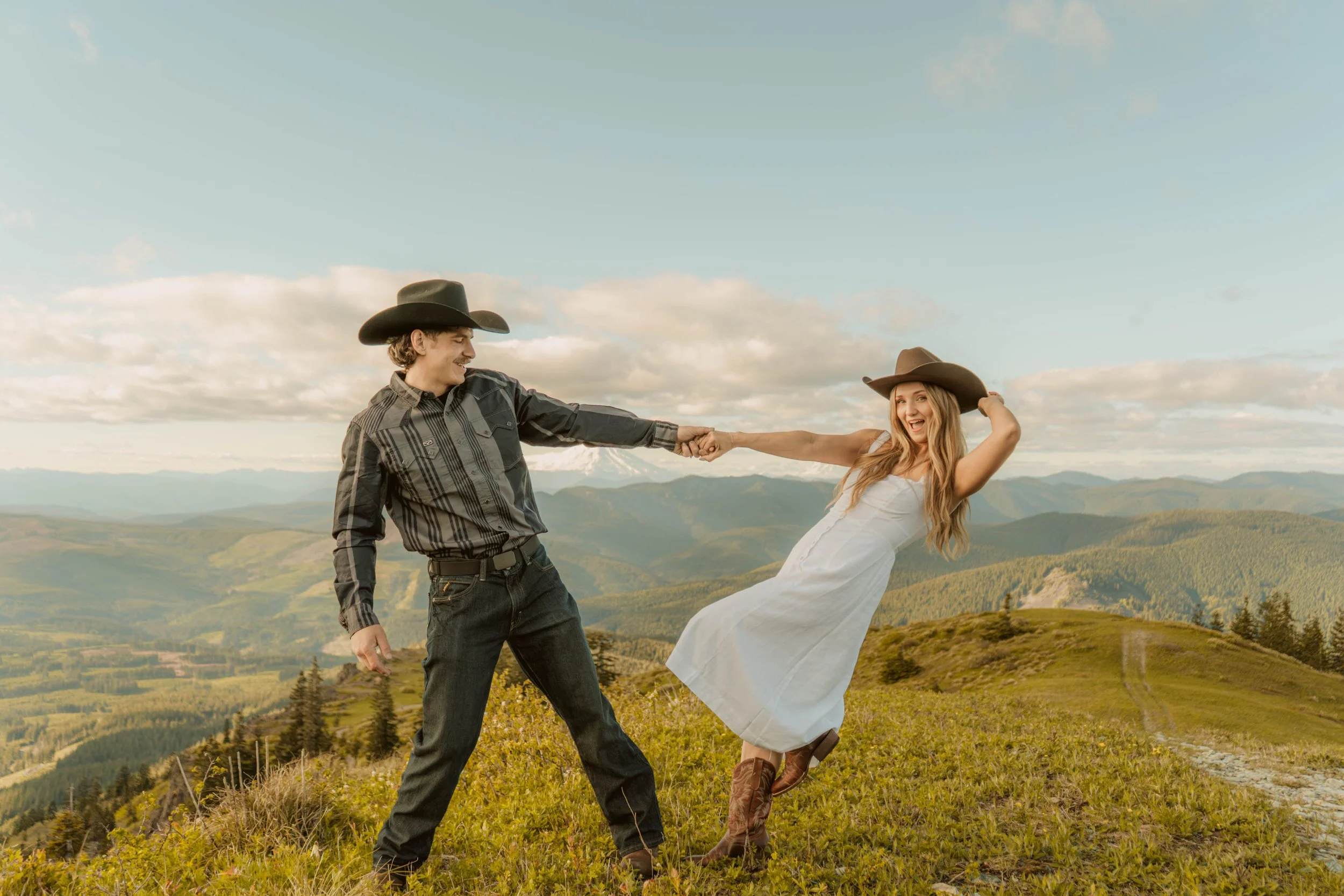 A young man and woman in cowboy hats and Western clothing dancing outdoors on a grassy hillside with rolling mountains and cloudy sky in the background.