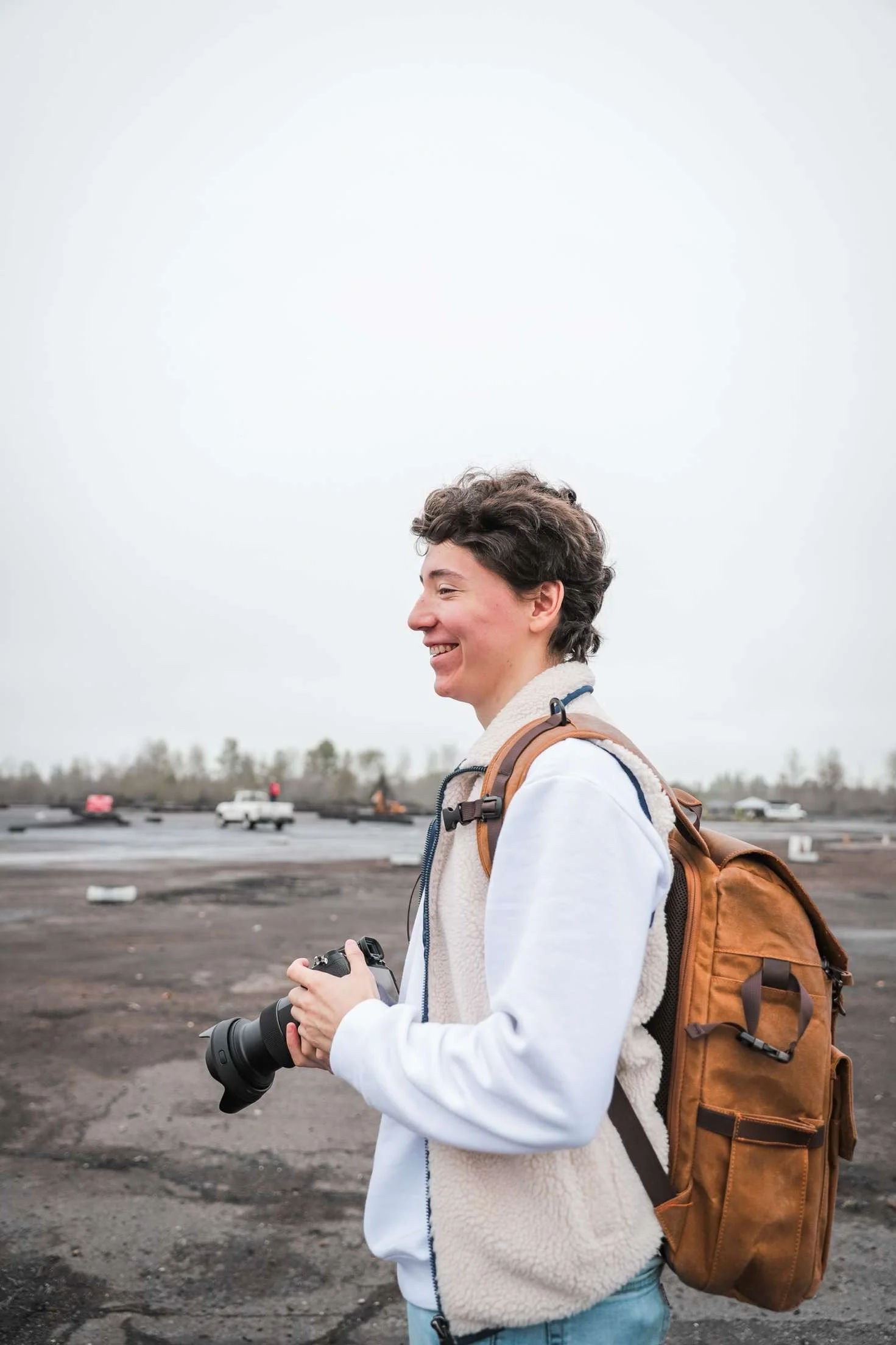 A young woman with short curly hair is standing outdoors on a cloudy day, smiling and holding a camera in her hands. She is wearing a white jacket with a beige fleece vest underneath and has a brown backpack on her back. The background shows a parking lot with some cars and a row of trees in the distance.