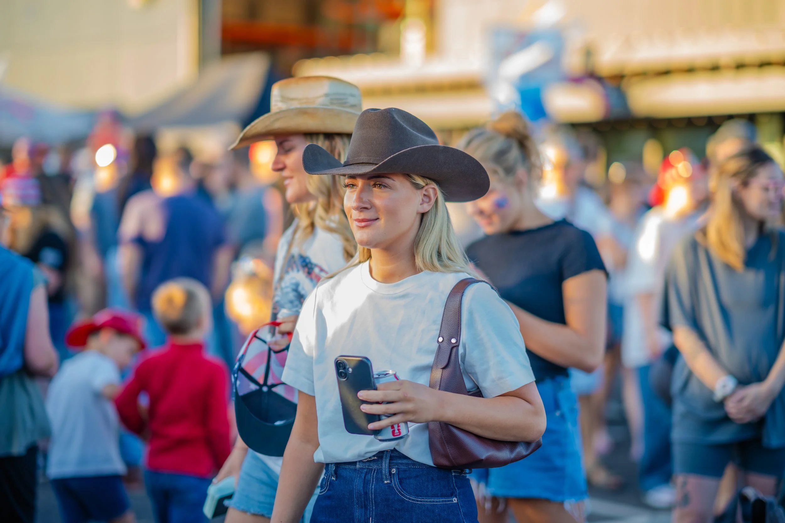 A young woman wearing a cowboy hat, white t-shirt, and carrying a purse and phone, stands amidst a crowd at an outdoor event during the evening. She is holding a drink and looking thoughtfully to the side.