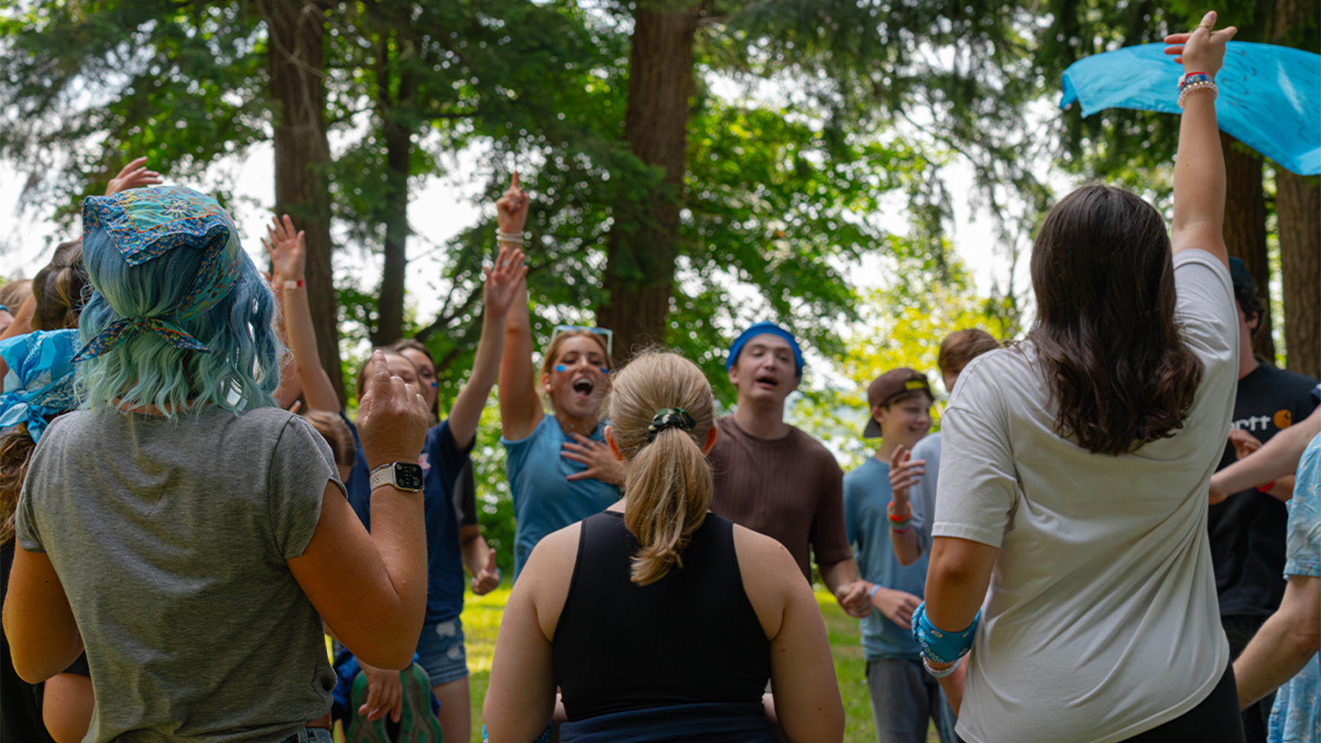 Group of people outdoors in a wooded area celebrating or having a joyful gathering, some with hands raised, wearing casual clothes and accessories.