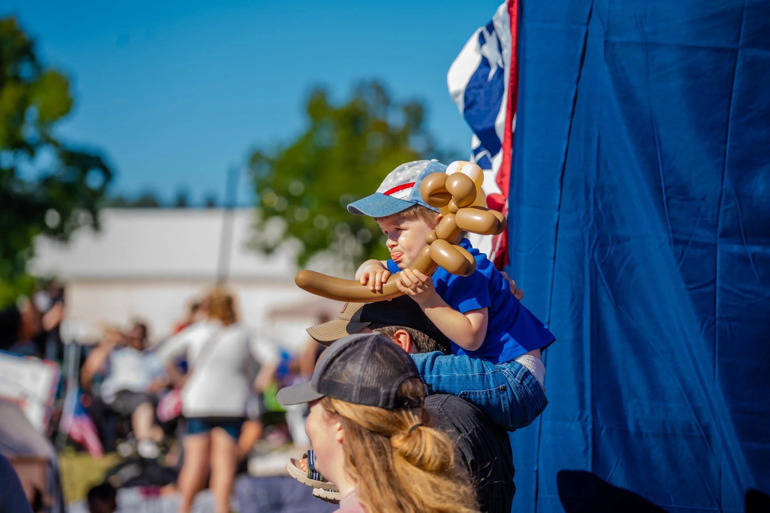 A young boy wearing a patriotic hat and blue shirt is sitting on an adult's lap at an outdoor event, holding a balloon animal. The scene is sunny with a crowd in the background.