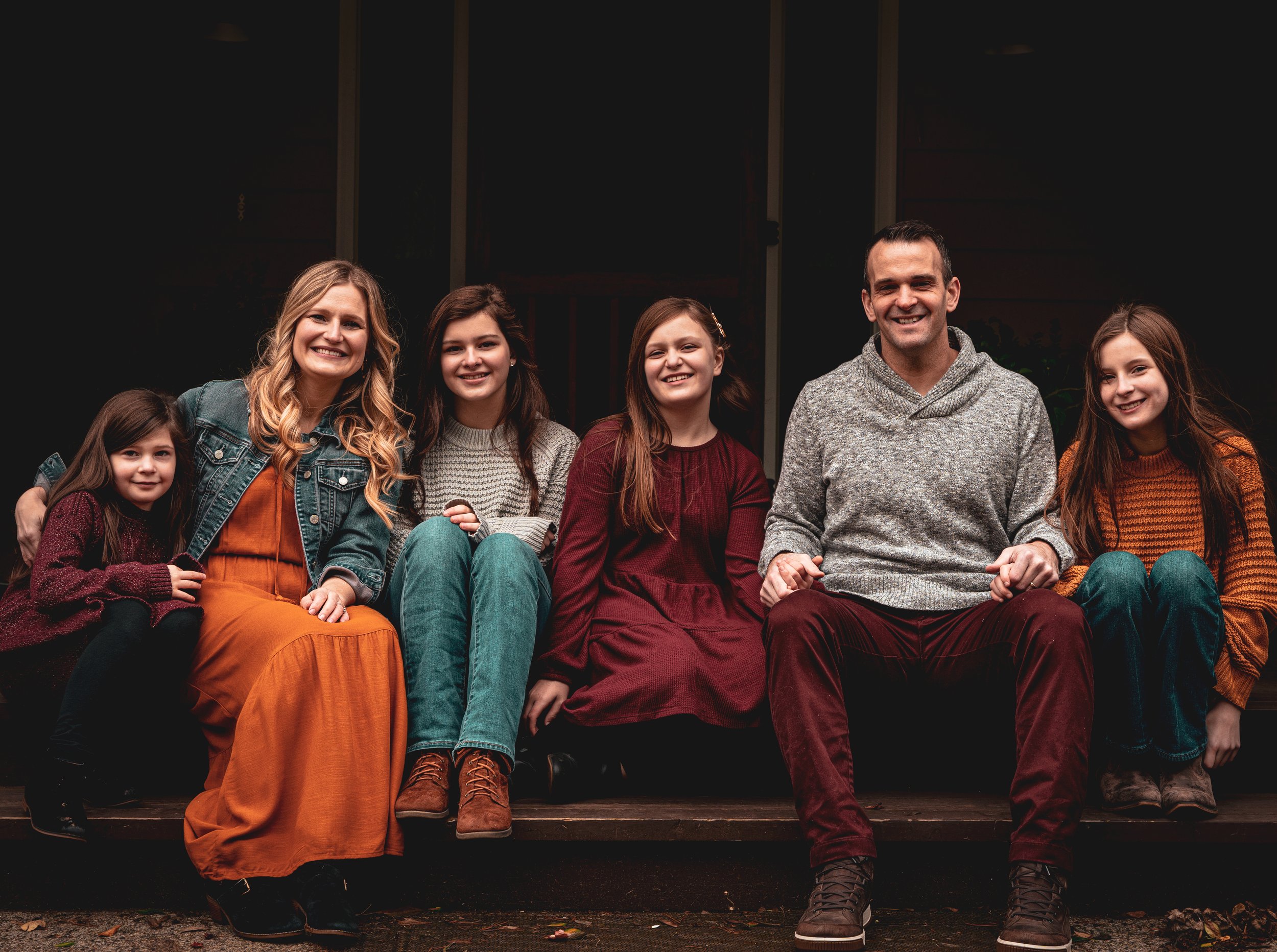 Group of six people, four children and two adults, sitting together on stairs outdoors, smiling at the camera during daytime.