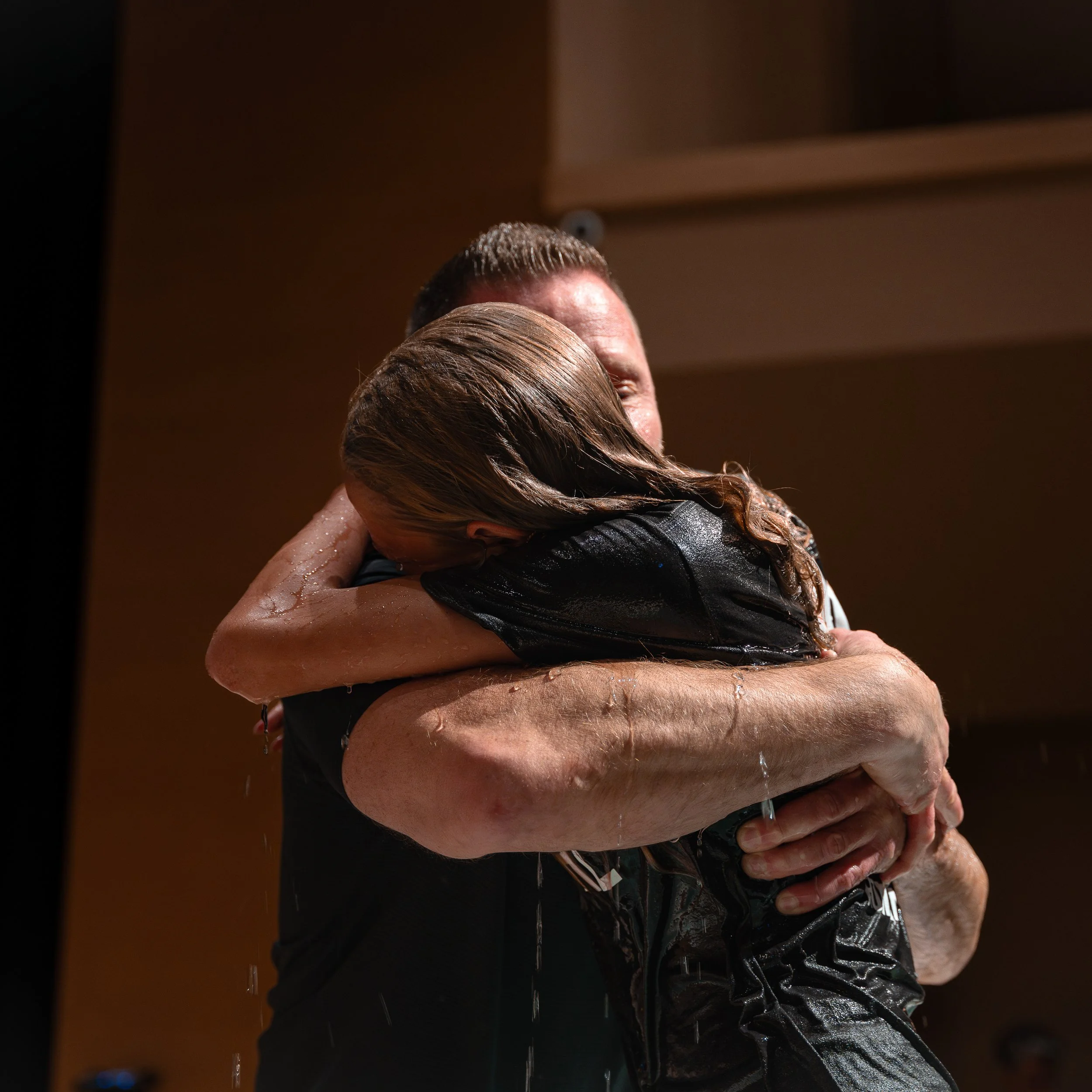 Two people embracing closely, with one person's head resting on the other's shoulder, surrounded by water droplets, in a warm indoor setting.