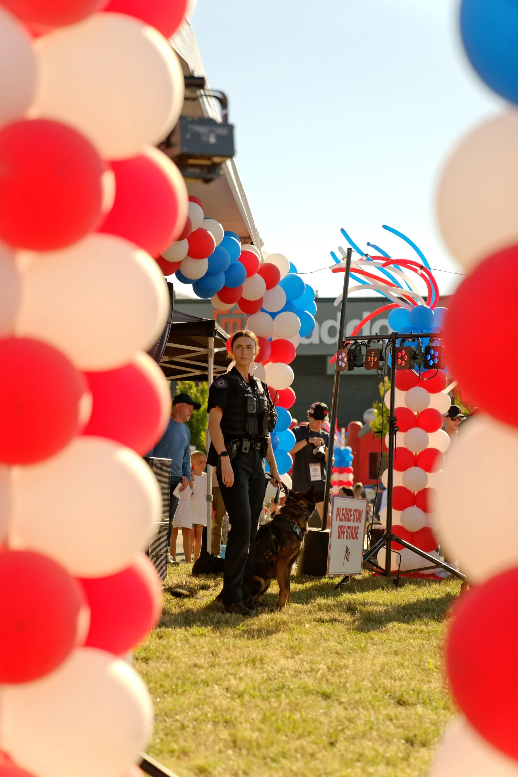 A parade or public event scene with balloons in red, white, and blue, and a police officer with a police dog among other people, tents, and decorations, under a clear sky.
