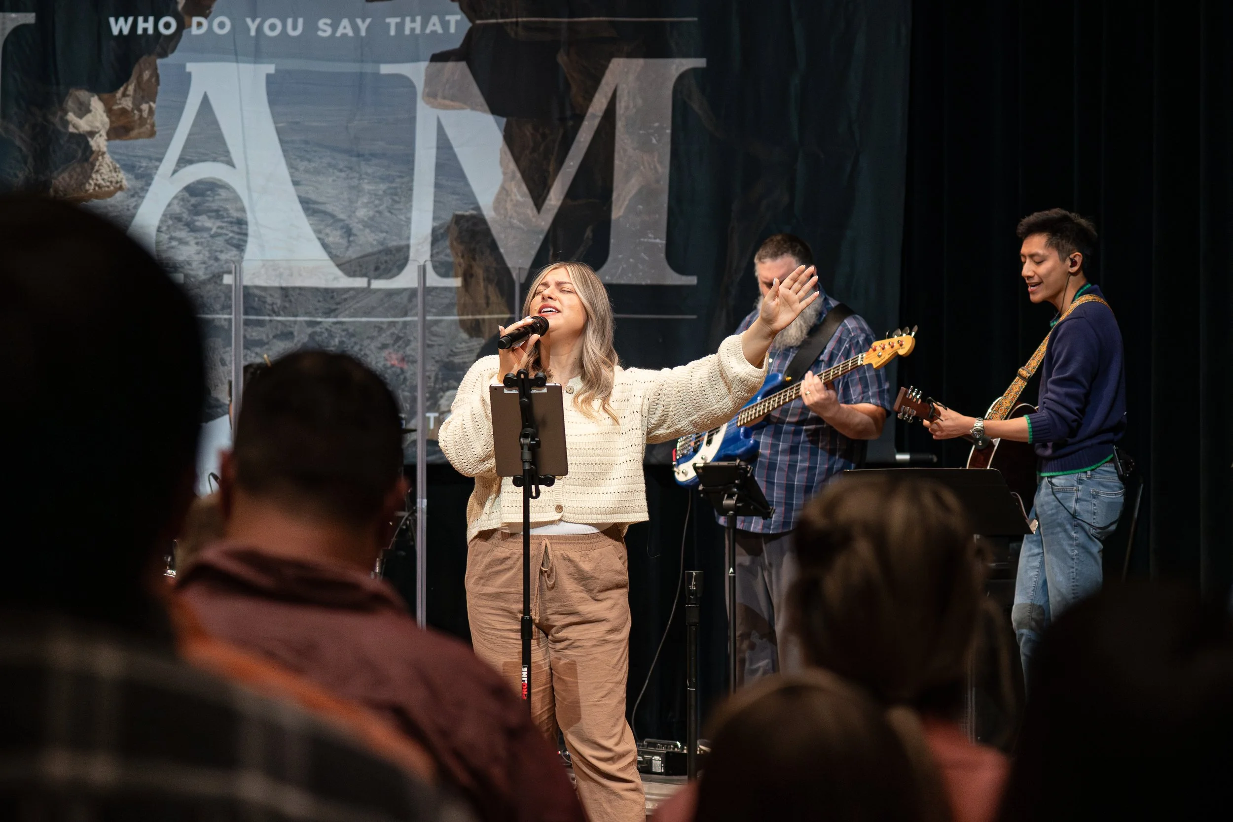 A woman singing into a microphone with her eyes closed and one arm raised, accompanied by two men playing guitars on a stage. There is a large backdrop with the text 'WHO DO YOU SAY THAT', partially obscured.