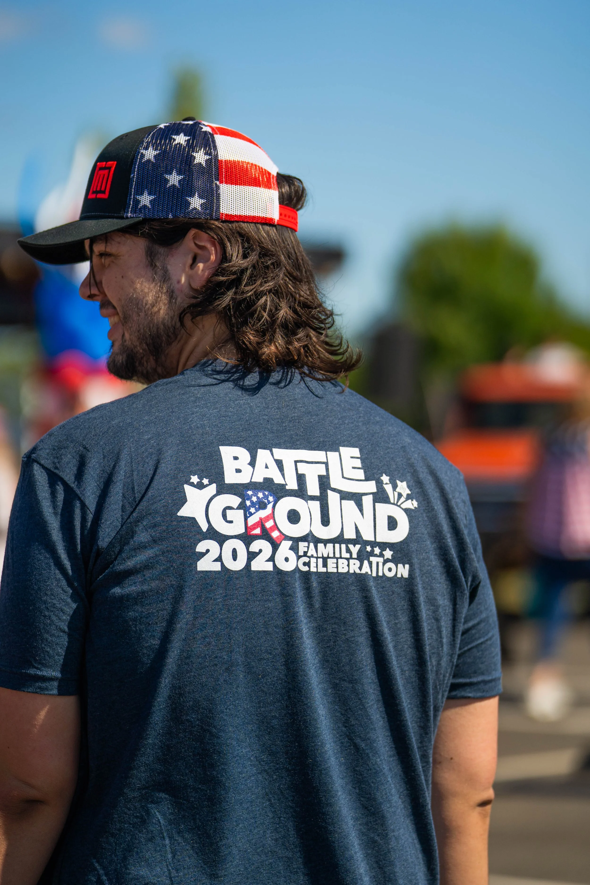 A man with long hair, beard, and mustache wearing a navy blue T-shirt with the words "Battle Ground 2026 Family Celebration" and a logo, and a baseball cap with an American flag pattern.