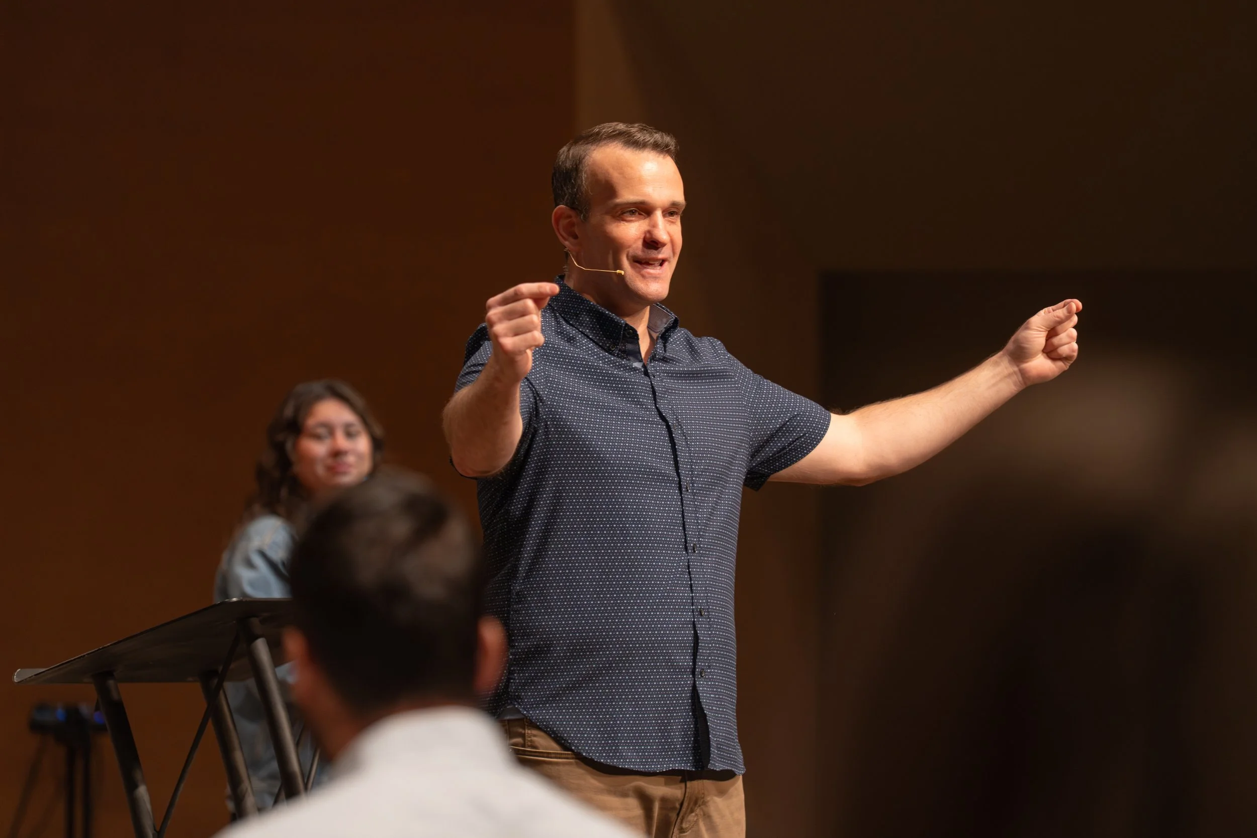 A man giving a presentation on stage with a microphone headset, gesturing with his hands, and an audience watching him.