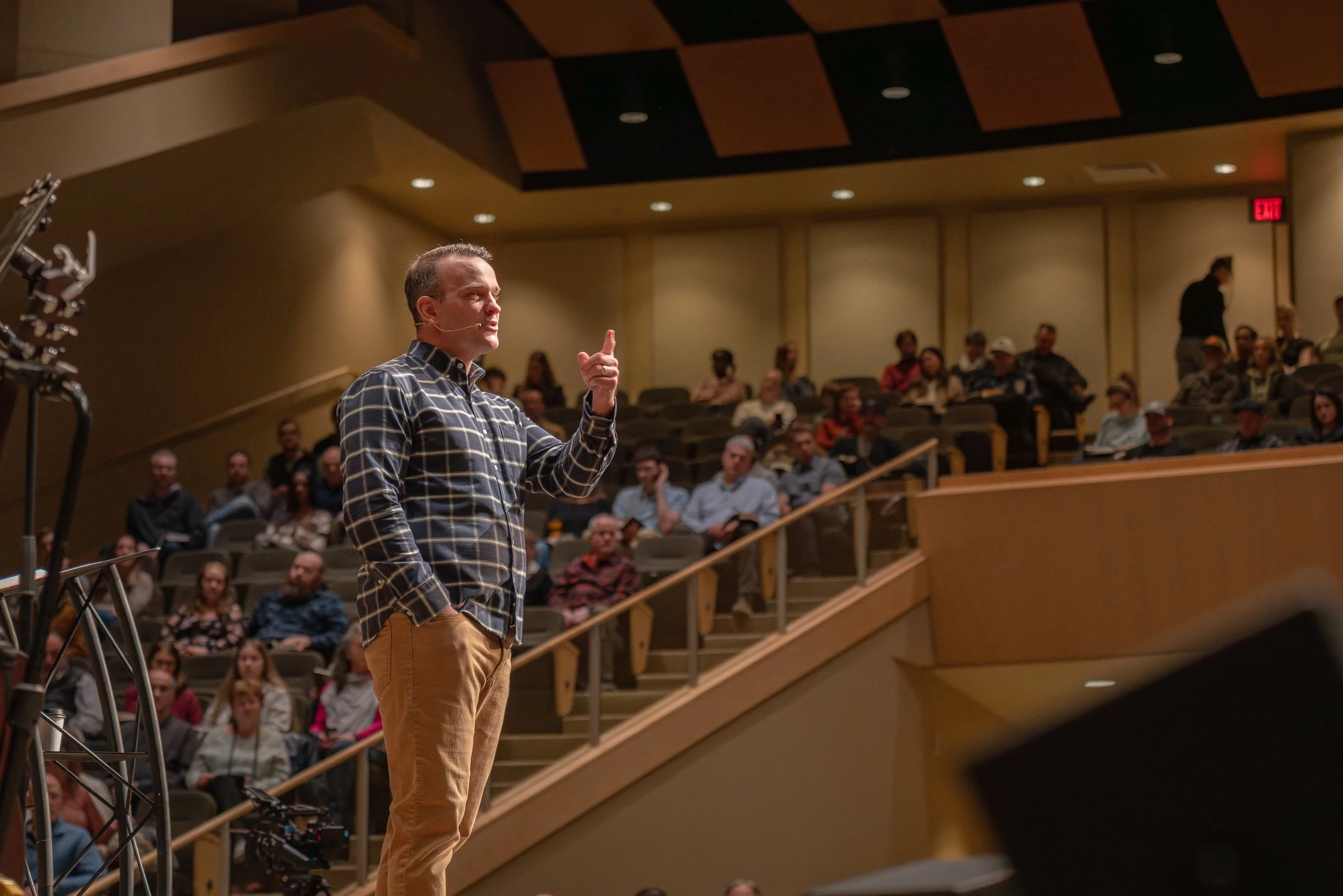A man in a plaid shirt speaking to an audience in a theater or lecture hall.