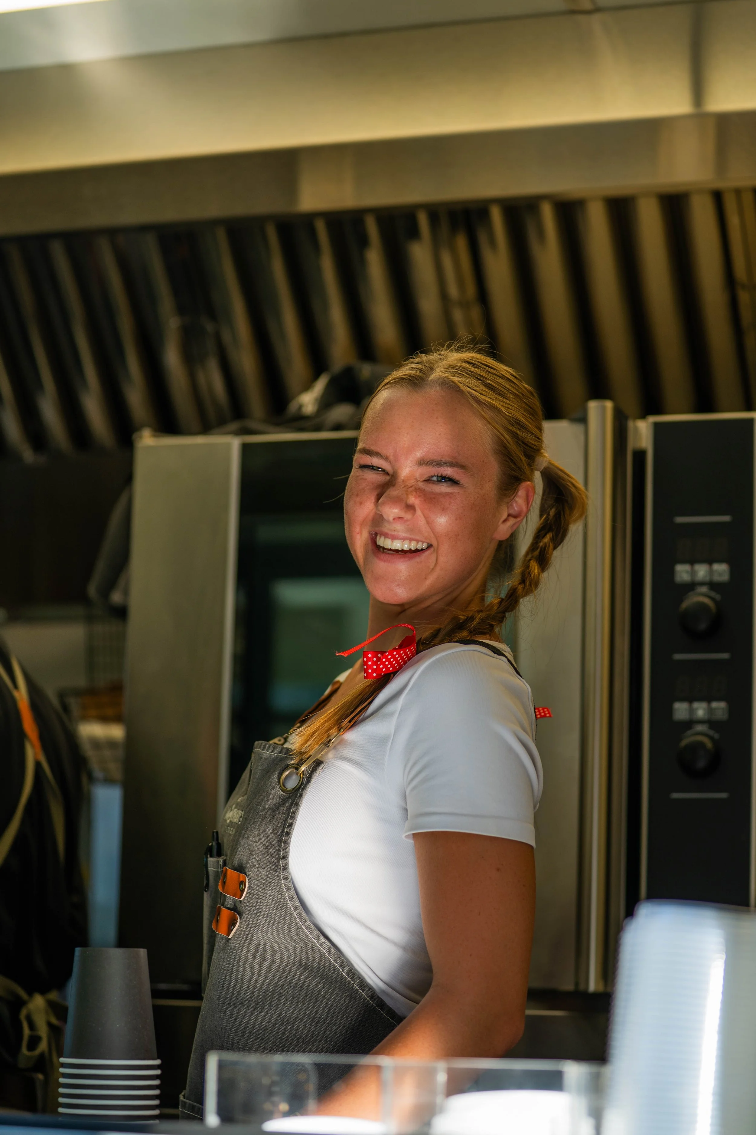 A young woman with red hair in a braid, wearing a gray apron with leather straps and a white shirt, smiling in a restaurant or cafe with a modern industrial design.