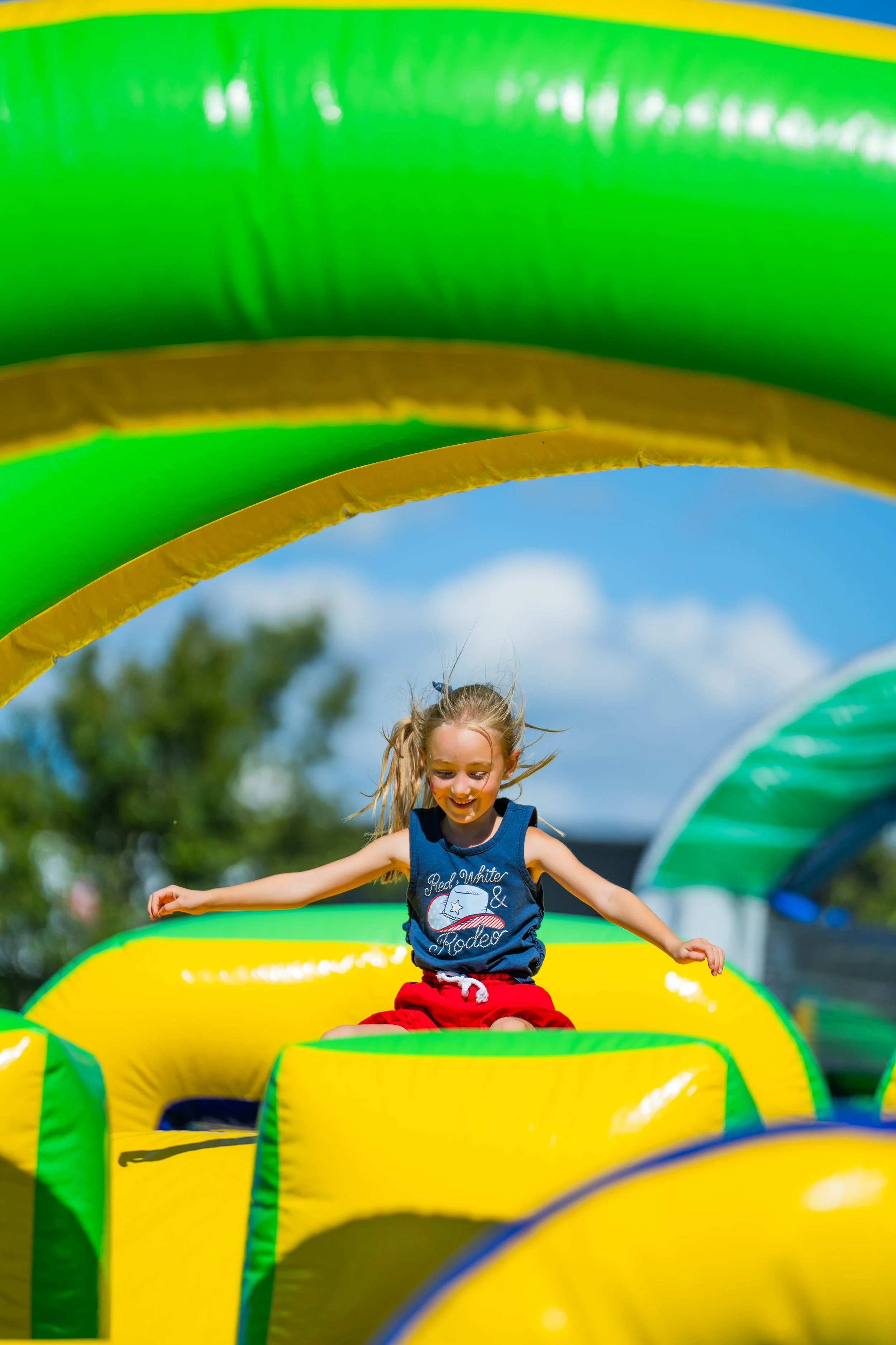 A young girl with blonde hair smiling as she plays on an inflatable obstacle course outdoors. The obstacle course has bright yellow, green, and blue sections. The girl is wearing a blue tank top with a patriotic design and red shorts.