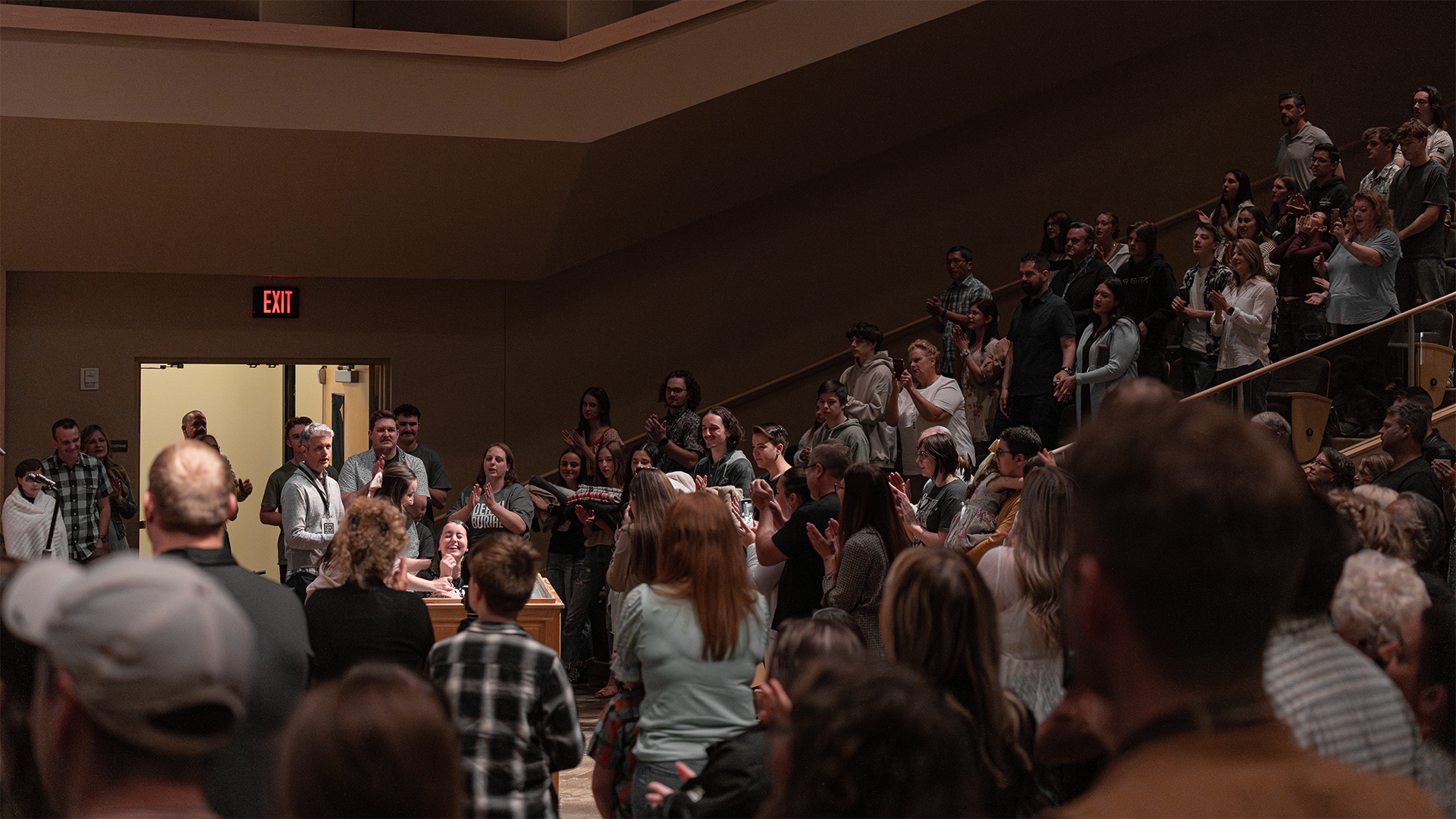 Crowd of people gathered in an auditorium, some standing and some seated, with an exit door visible in the background.