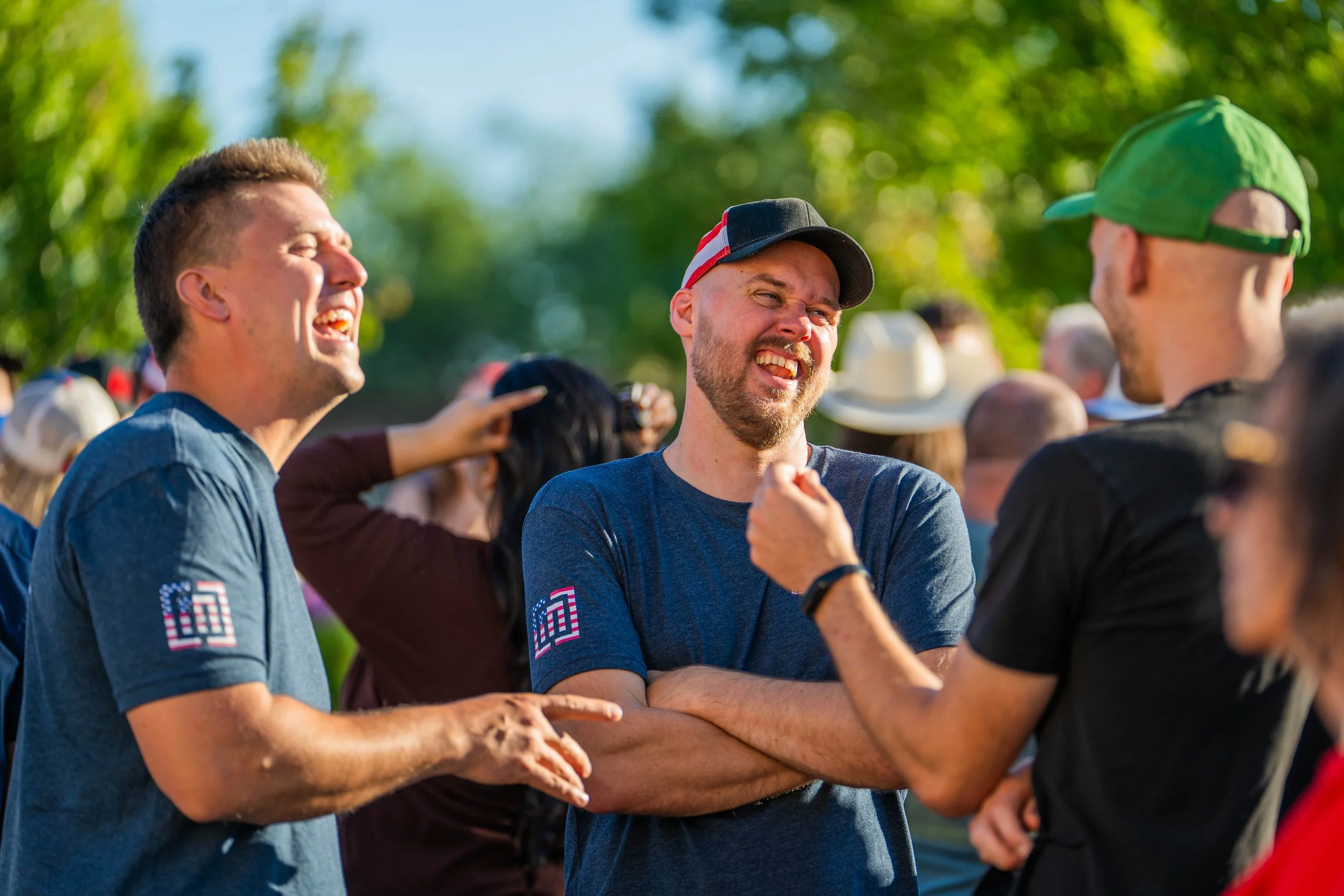 Three men are laughing and talking outdoors during daytime, surrounded by other people and green trees in the background.