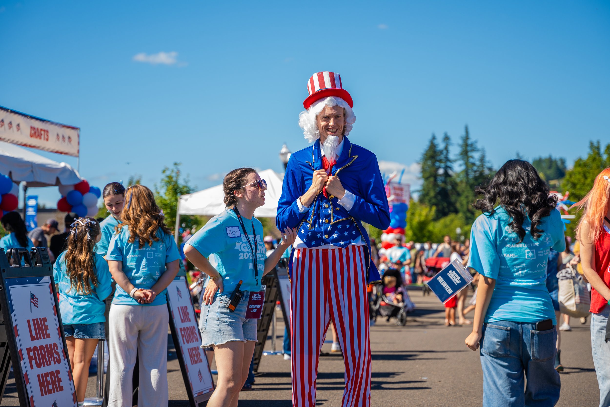 A man dressed as Uncle Sam with a white beard and red, white, and blue striped pants, standing among a group of people at an outdoor event on a sunny day. The man is smiling and holding a hand to his chest.