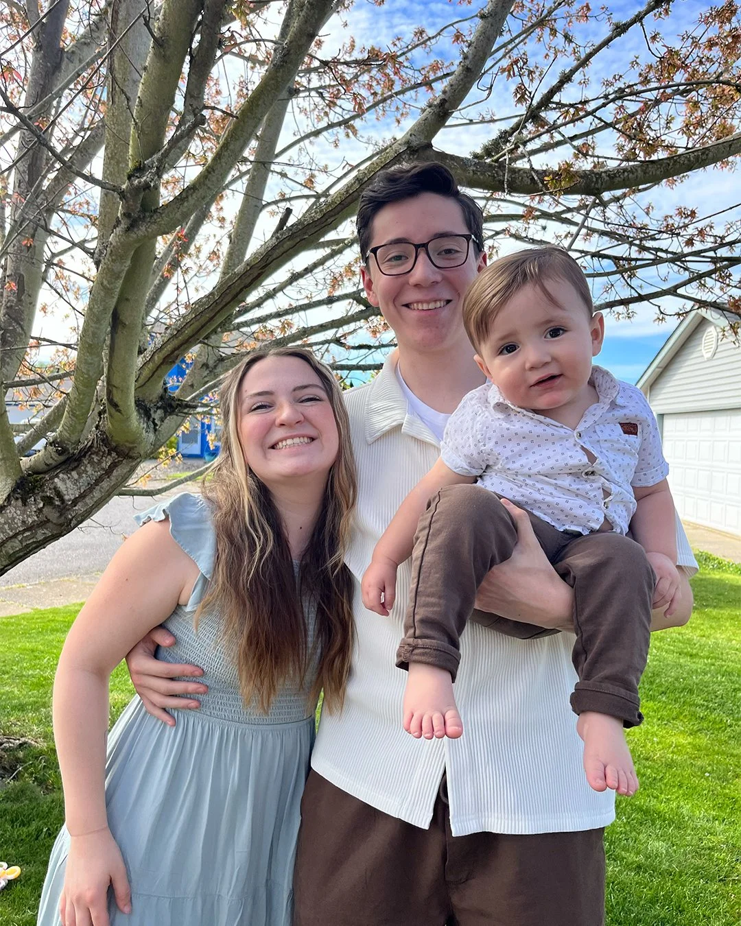 A happy family of three standing outside under a tree with budding leaves, with a house and a garage in the background. The woman is smiling with her arm around the man, who is holding a young boy. The boy, sitting on the man's arm, is looking at the camera.