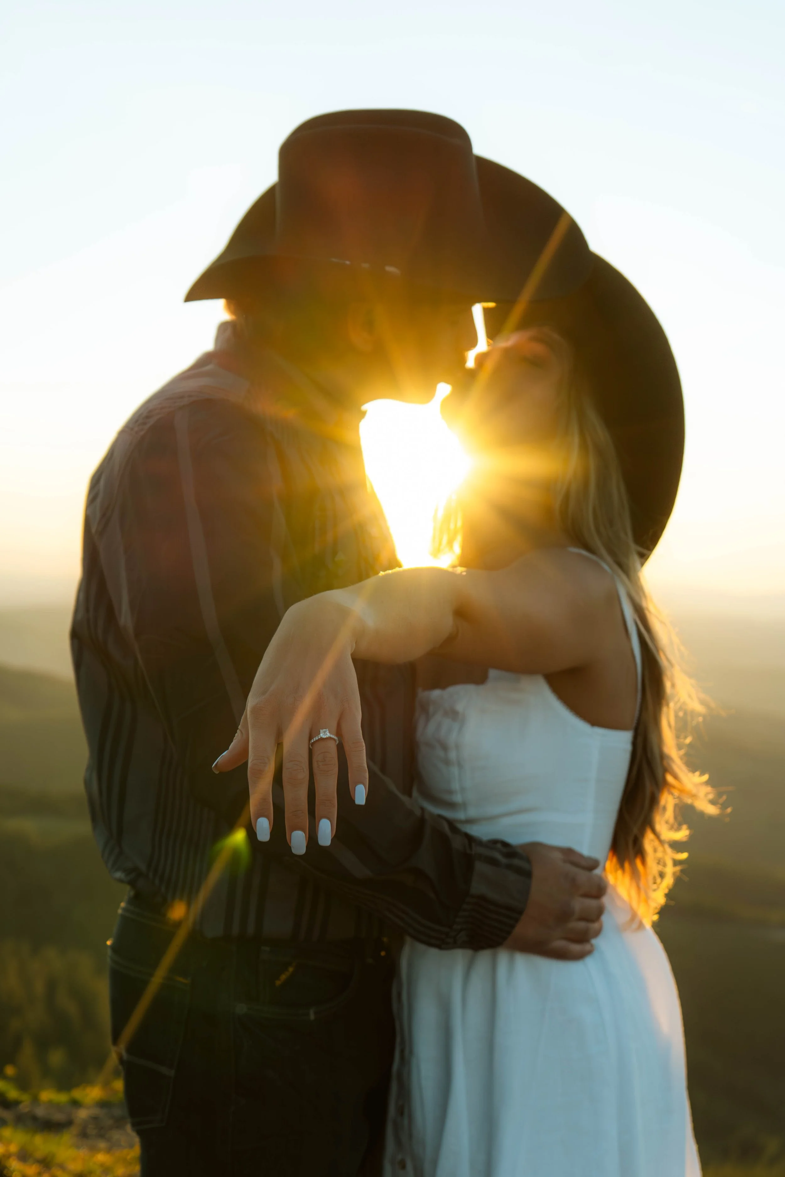 A couple kissing during sunset, the woman with a black wide-brimmed hat and a white dress, the man with a cowboy hat and plaid shirt, in an outdoor setting with a blurred background.