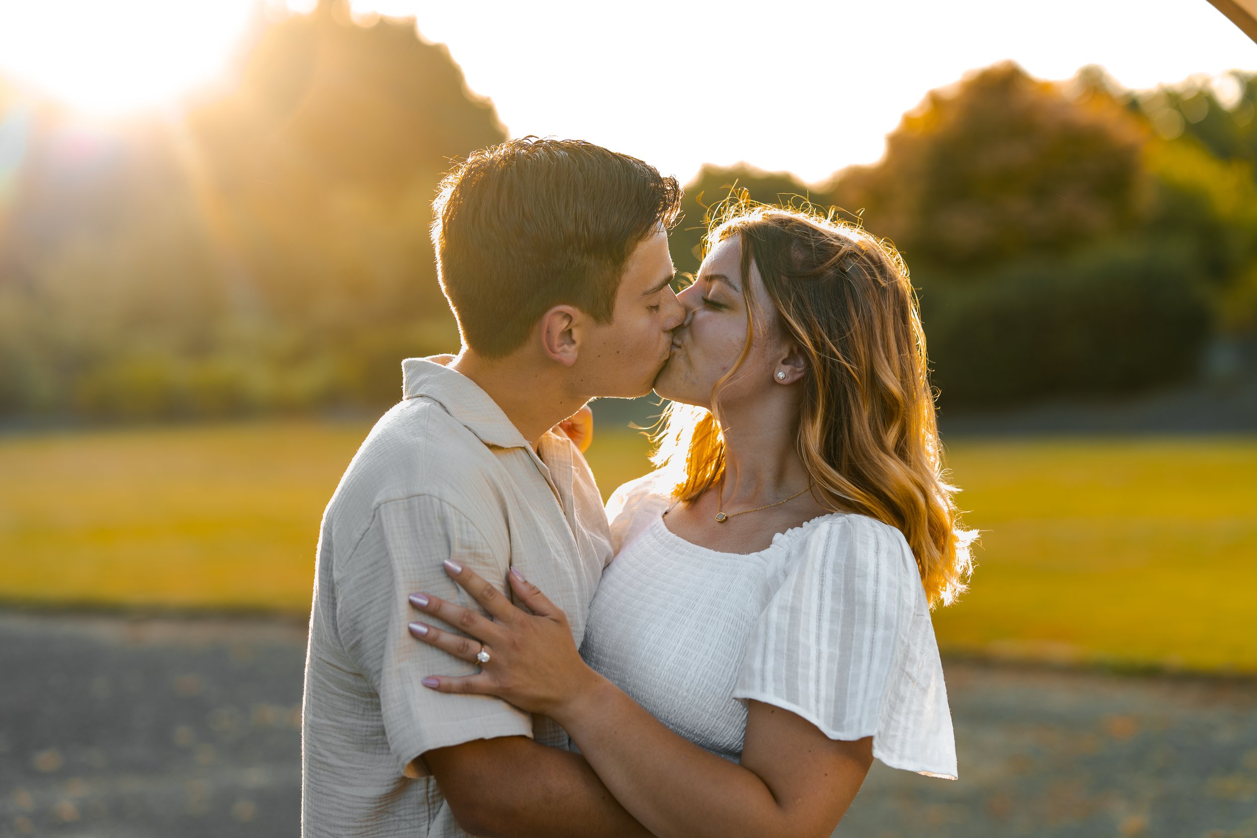 A young couple kissing outdoors in the late afternoon sun, with trees and a blurred background.